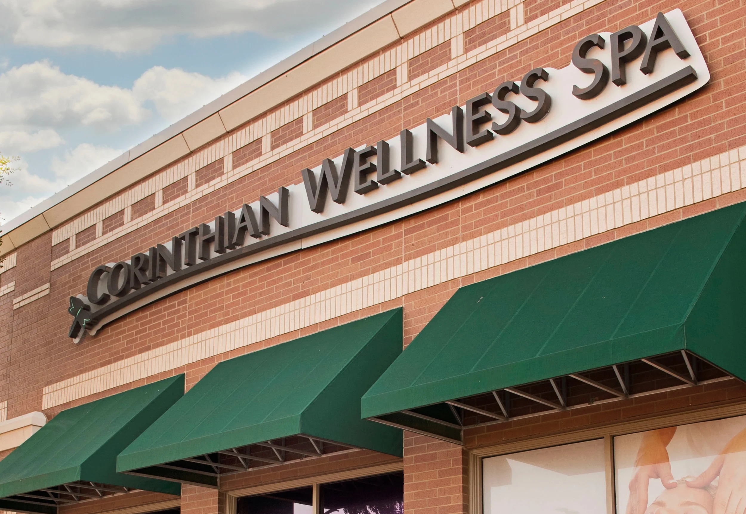 Exterior of a wellness spa with a brick facade, green awnings, and a sign reading 'Corthian Wellness Spa' against a cloudy sky.