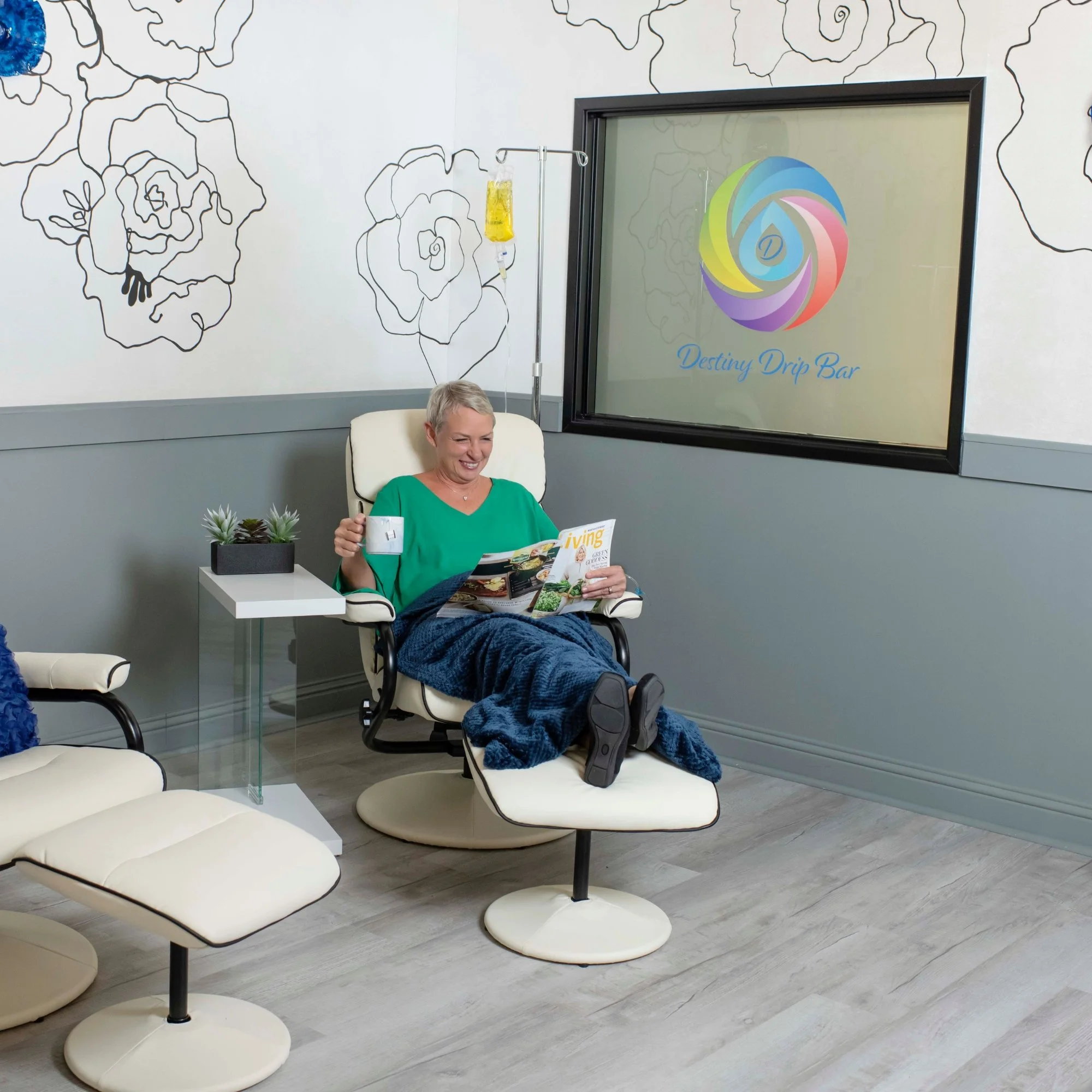 A woman sitting in a white chair, smiling and reading a magazine titled "Living", in a modern waiting room with white and black chairs, a small table with a plant, and a perforated wall mural of roses.