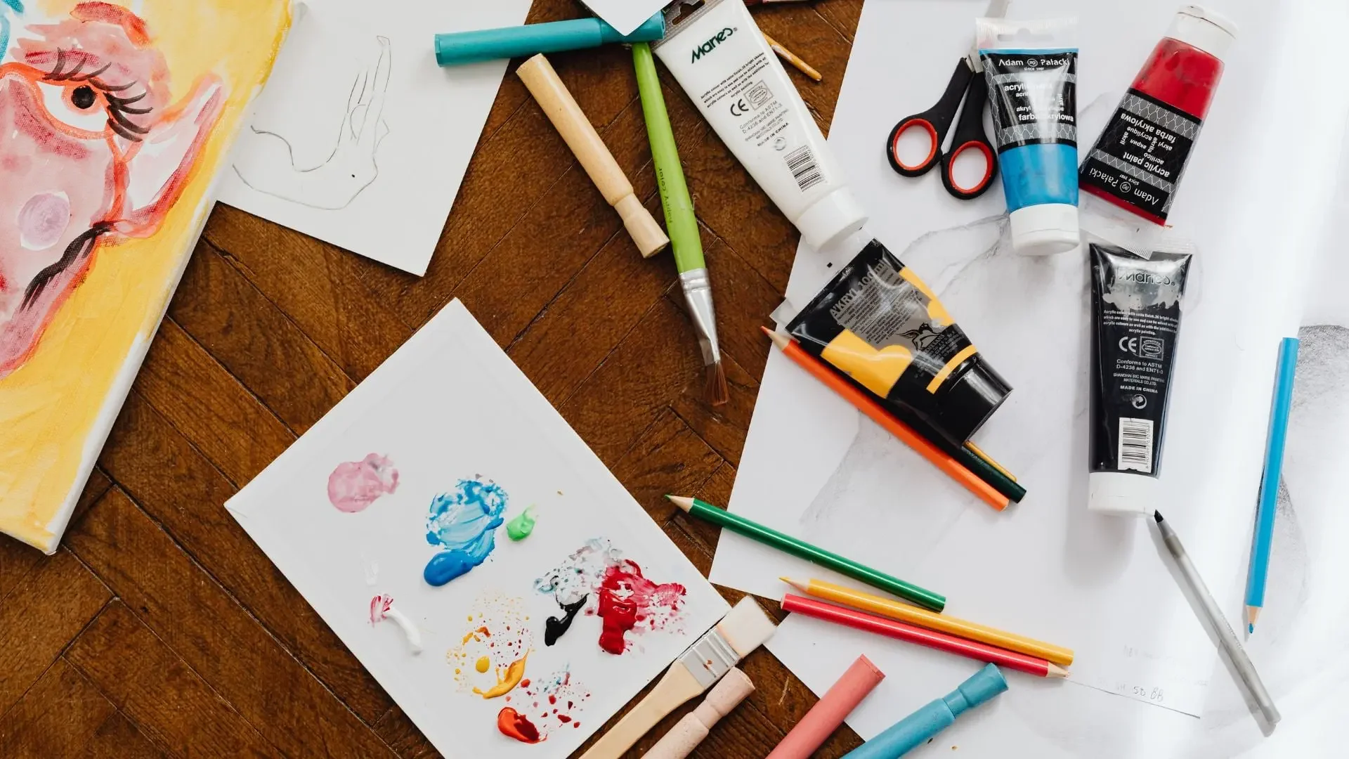 Art supplies scattered on a wooden floor, including tubes of paint, brushes, colored pencils, scissors, and a partially completed canvas painting of an abstract face.