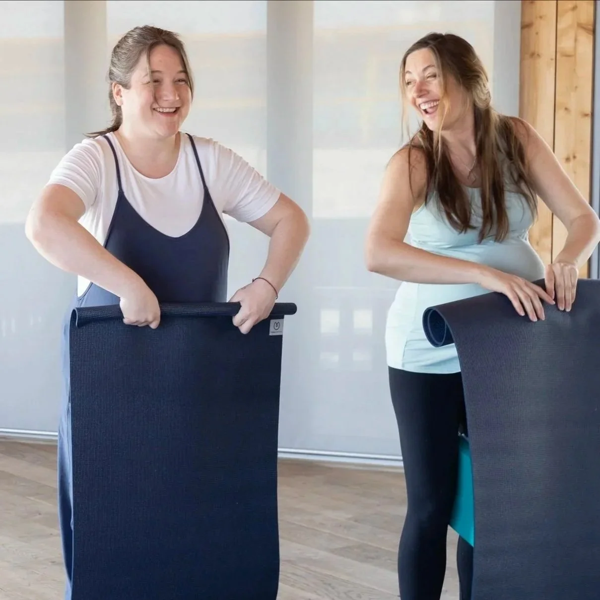 Two women are standing next to each other, laughing, and holding yoga mats in a bright indoor space.
