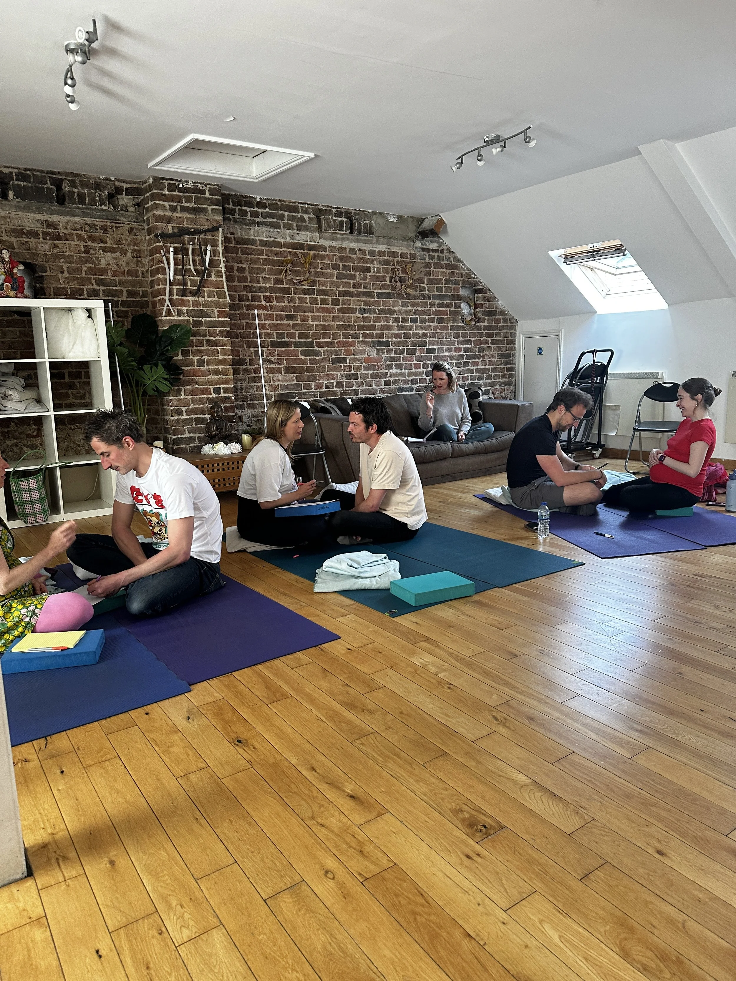 Group of people participating in a yoga or meditation class in a bright, wooden-floored studio with brick and white walls, some sitting on mats and some on a sofa, engaging in conversation or meditation.