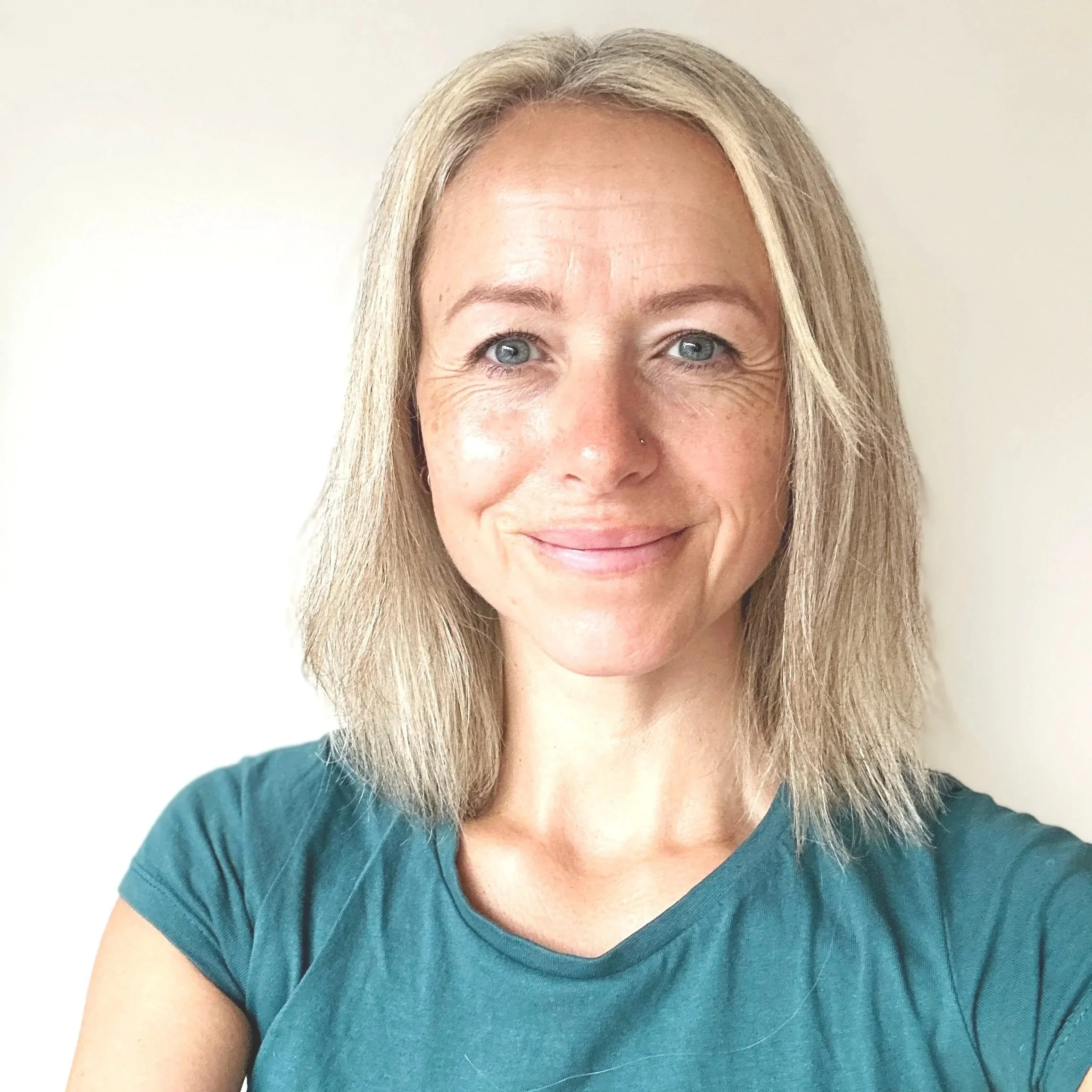 A woman with shoulder-length blonde hair, blue eyes, and a small nose piercing, smiling at the camera against a plain white background, wearing a teal T-shirt.