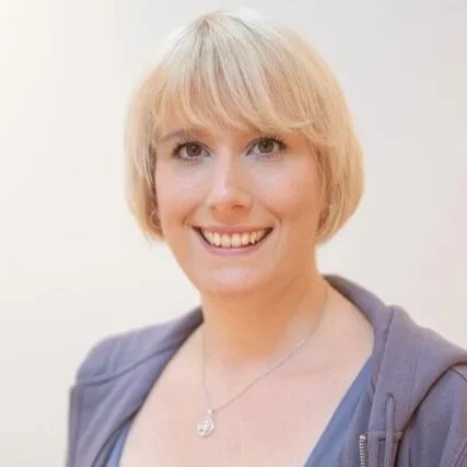 A woman with short blonde hair smiling, wearing a blue top and a gray hoodie, standing against a plain white background.