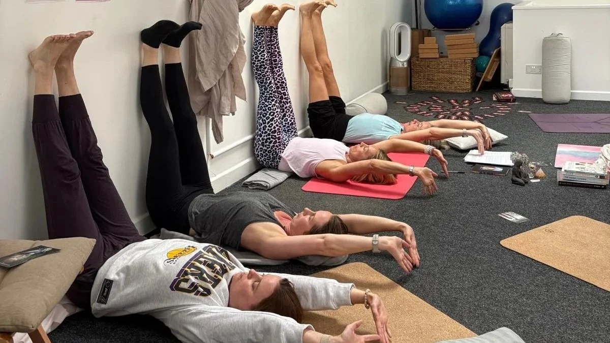 Four women practicing yoga in a studio, lying on their backs against the wall with legs raised vertically and arms extended forward.