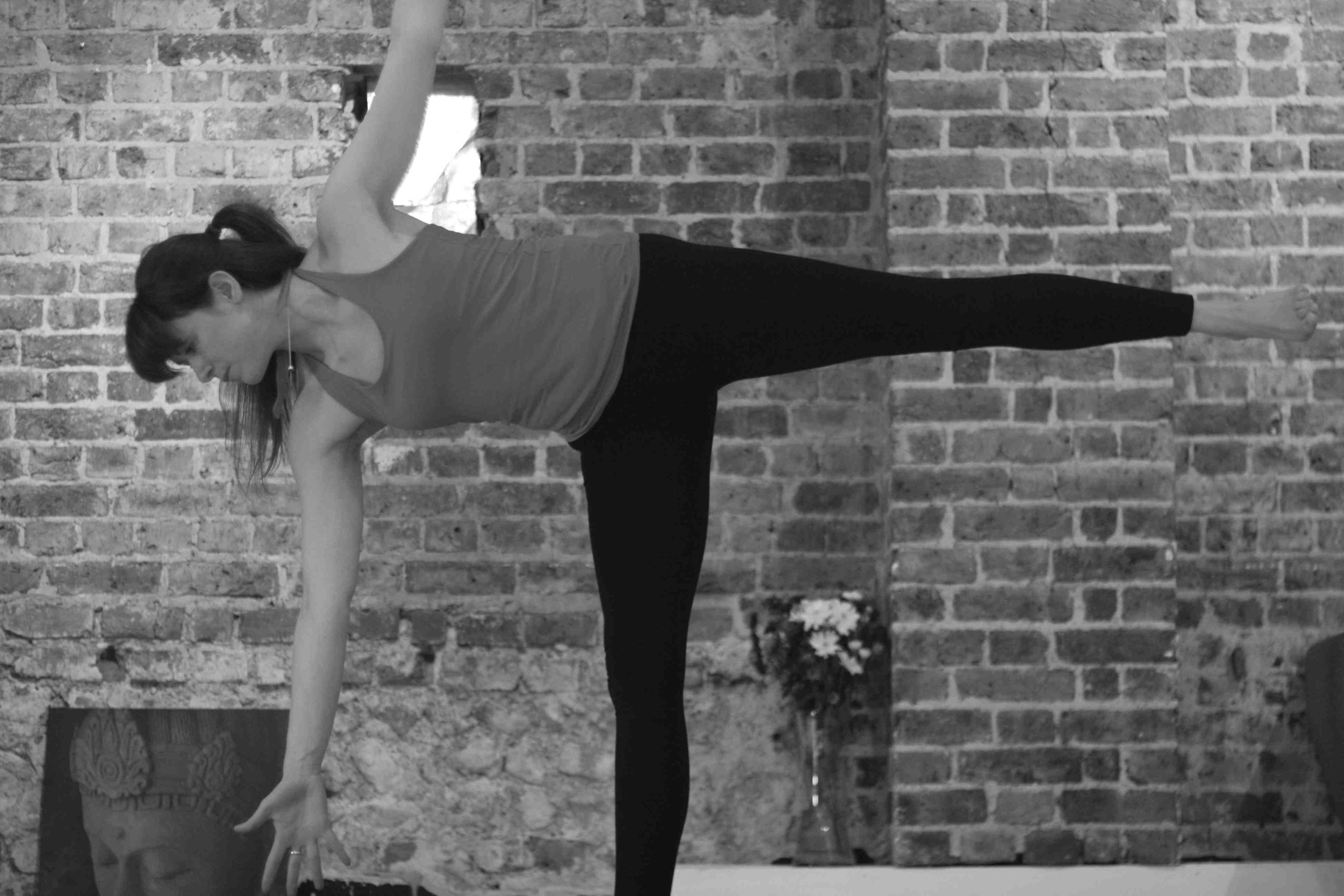 A person practicing yoga indoors against a brick wall. The person is in a standing forward bend pose, reaching one hand toward the floor while the other arm is extended upward. The individual is wearing a sleeveless top and leggings.