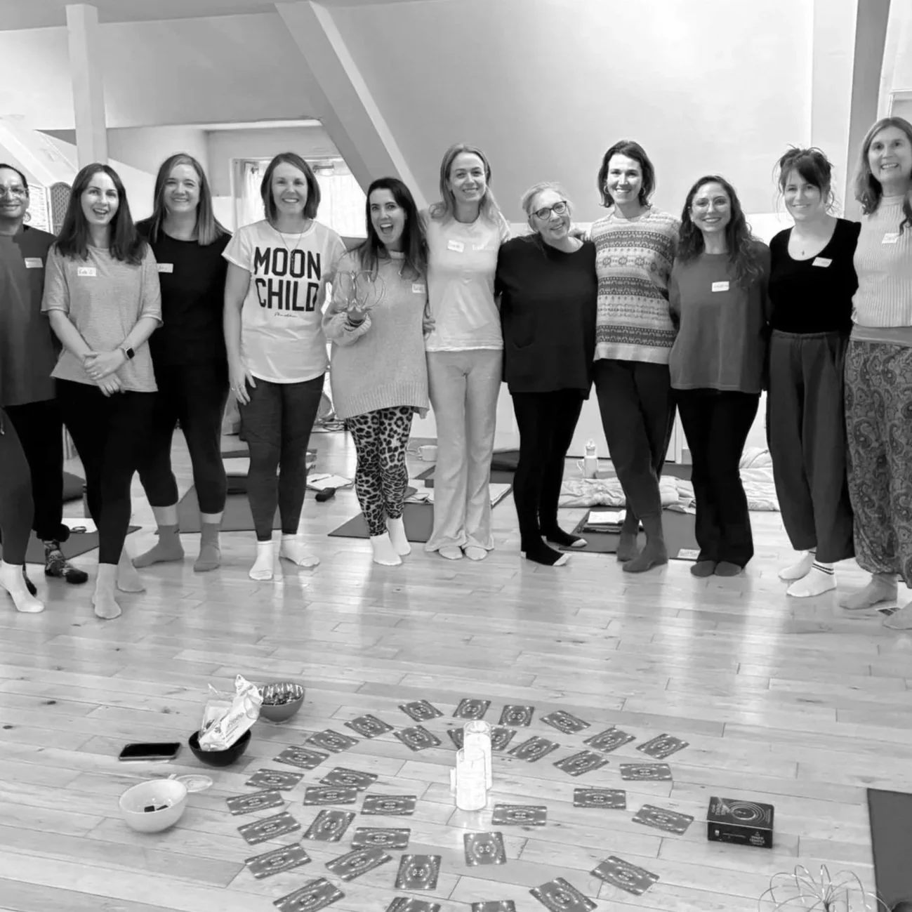 A group of eleven women standing together indoors, smiling at the camera during a gathering or celebration. The floor has cards arranged in a circle, with bowls and bottles nearby.