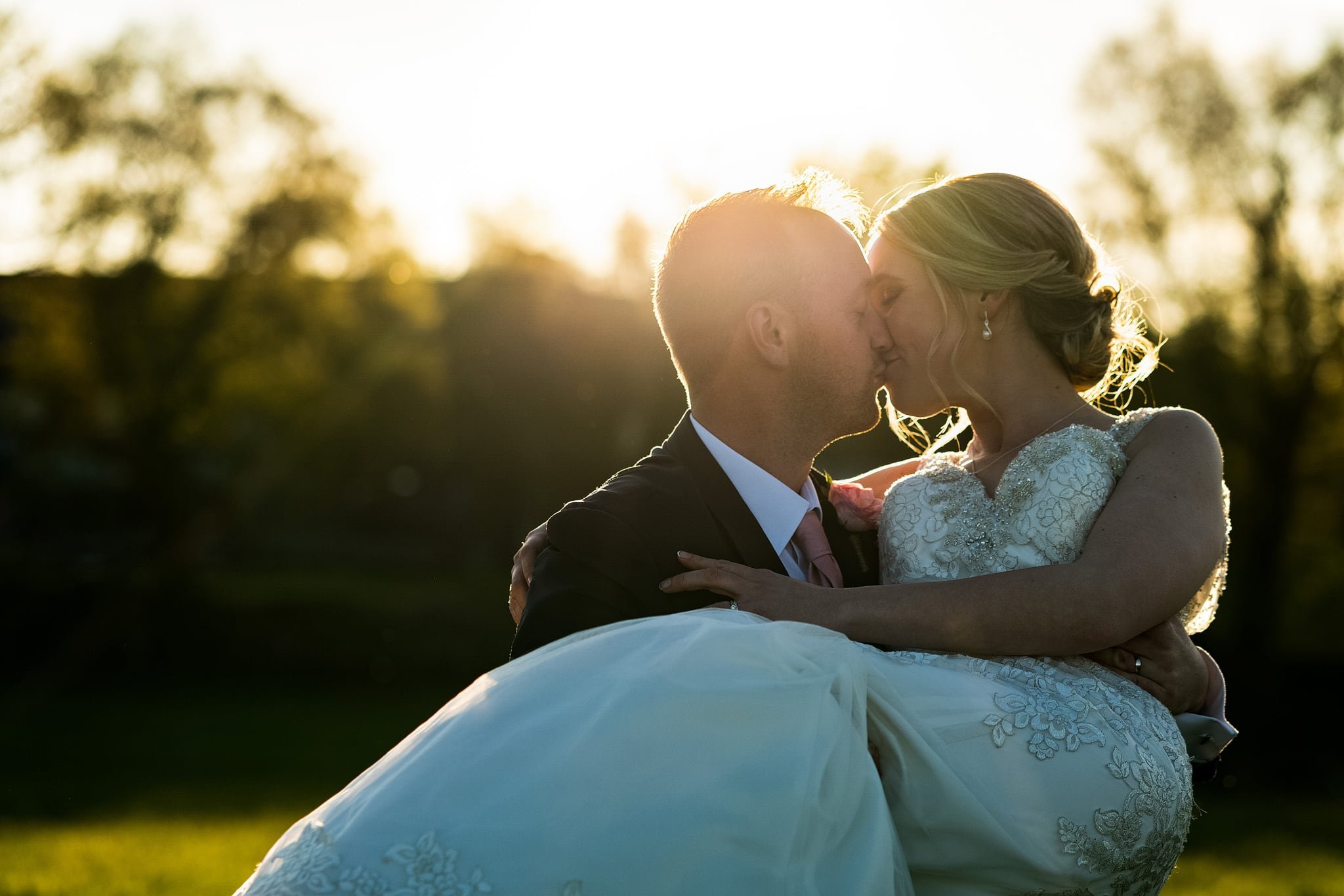 Groom kissing bride whilst carrying her at sunset