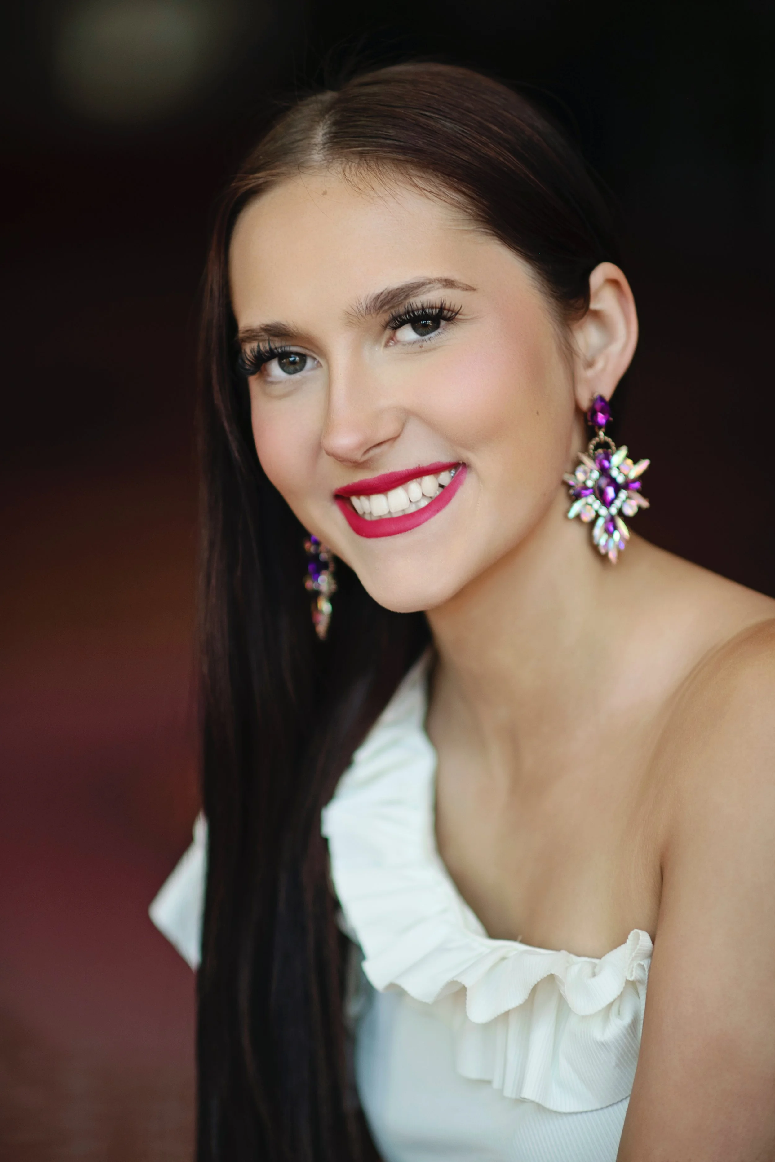 A young woman with brown hair, braces, and a bright smile, wearing a cream-colored ribbed top and a delicate gold necklace, standing against a plain light background.