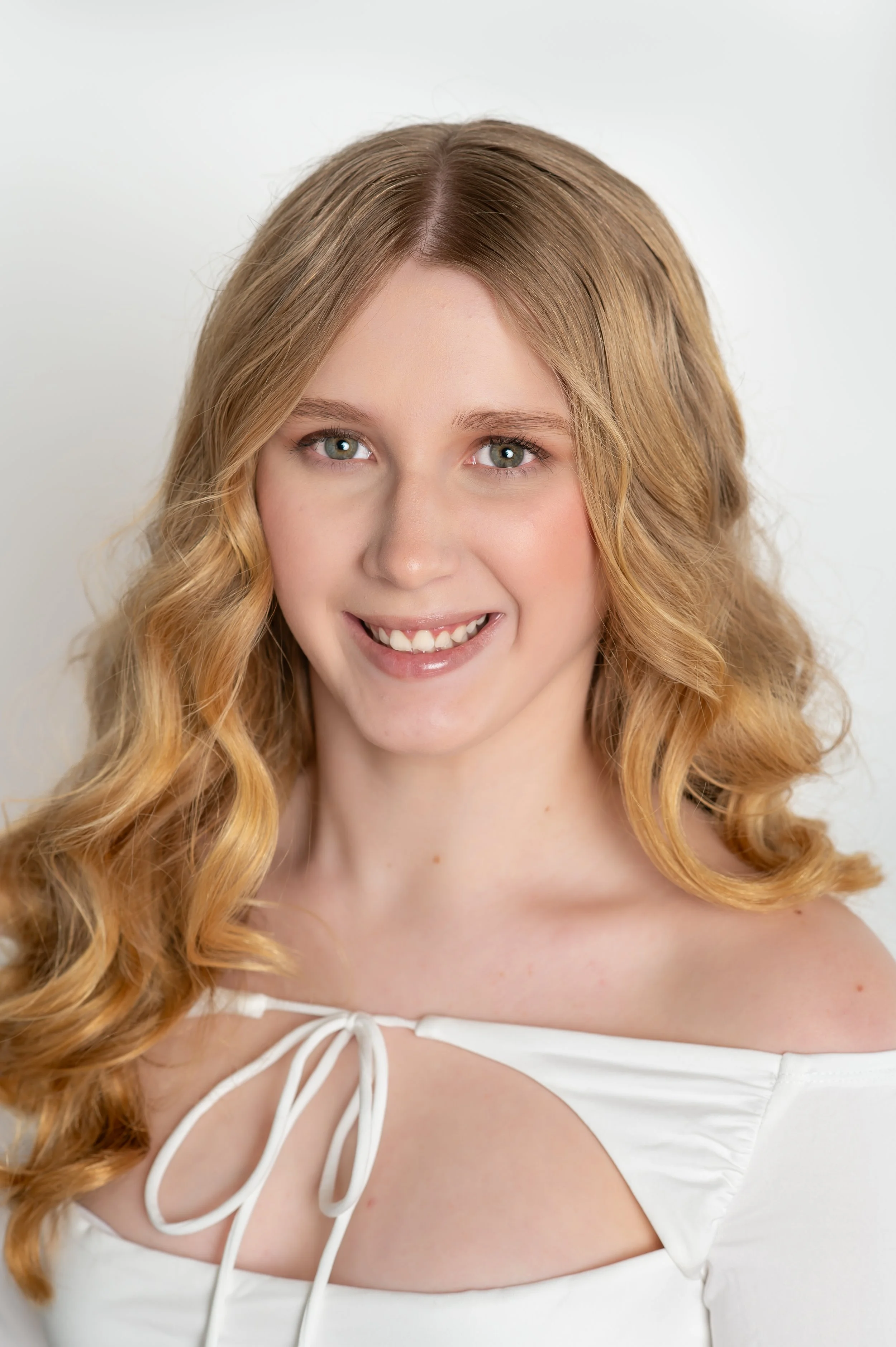A young woman with long wavy brown hair, smiling, wearing a white top with sheer sleeves adorned with 3D floral appliques, standing against a plain light gray background.