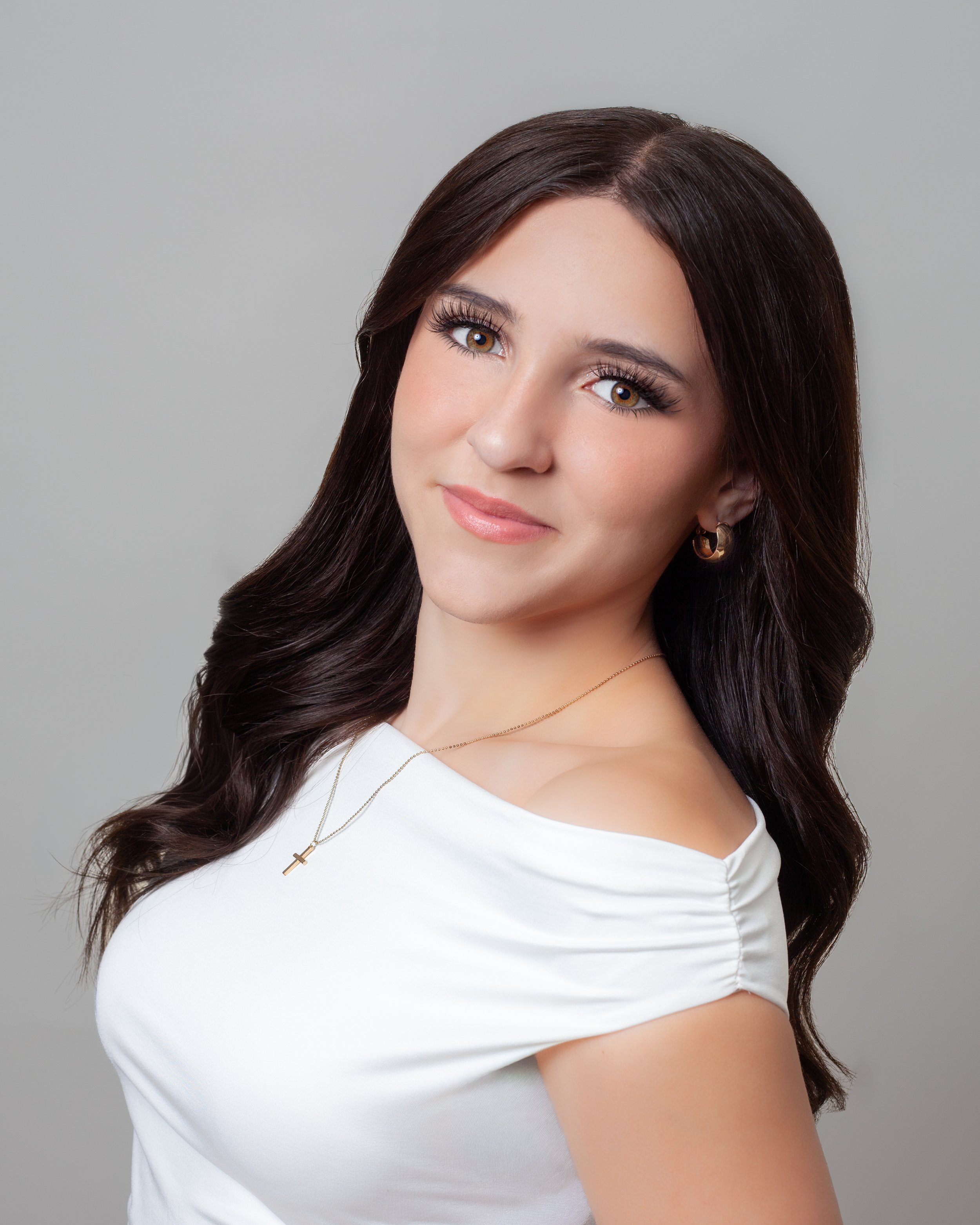 Young woman smiling with long brown hair and wearing a white top, posing with her hand on her chin against a plain background.
