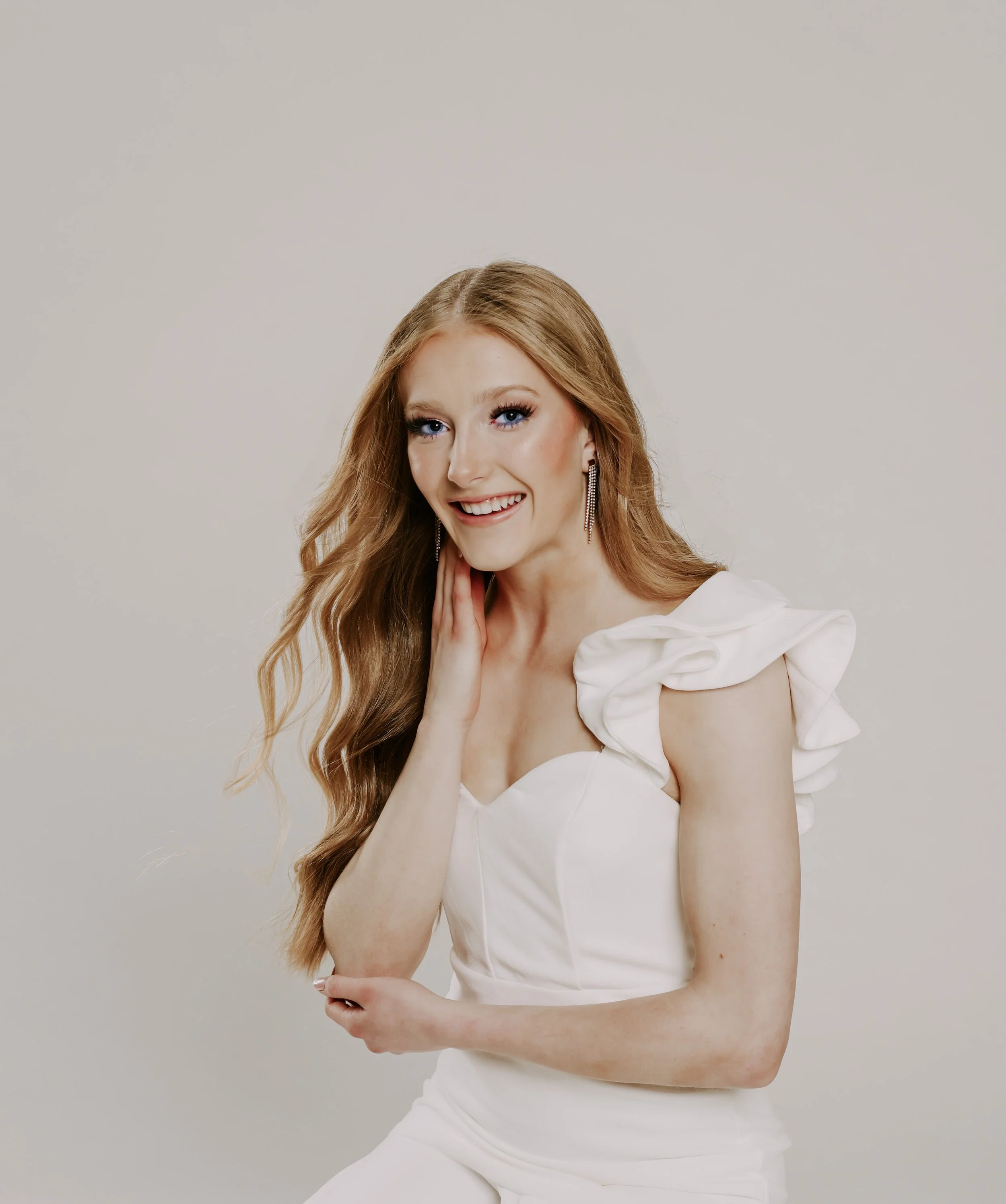 A person with long blonde hair wearing a white blouse and smiling, with a hand near the face and wearing teardrop earrings against a plain background.