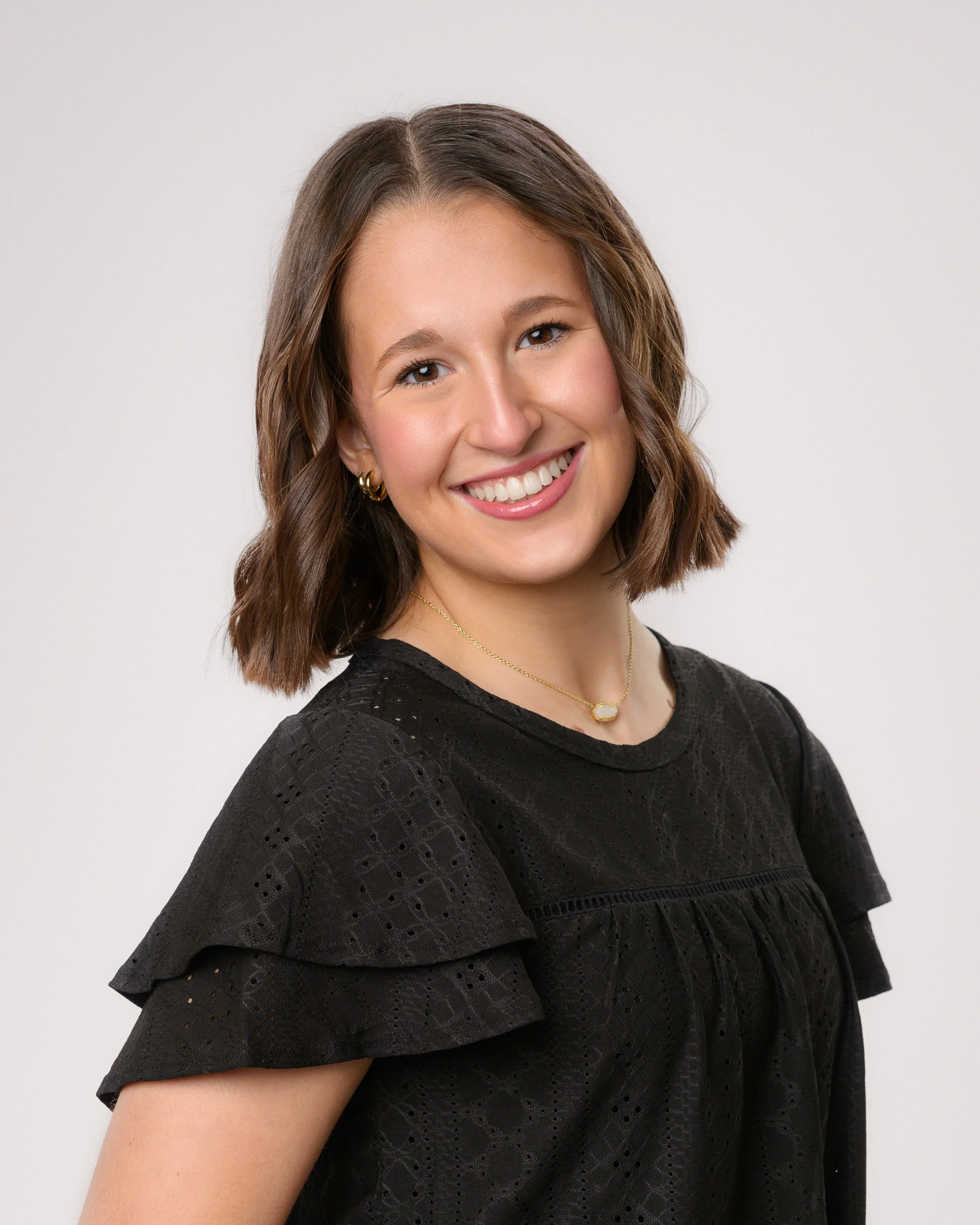 Smiling woman with long wavy hair wearing a black strapless dress and sparkling earrings, posing against a light background.