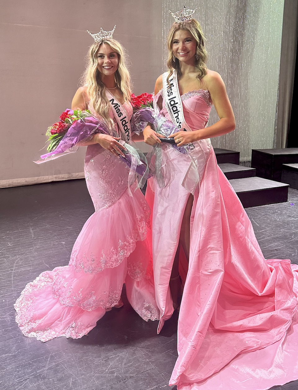 Two women wearing sashes and crowns, one in a lavender gown and the other in a green gown, standing together at a beauty pageant event with pink and gold balloon decorations and a large illuminated 'LOVE' sign in the background.
