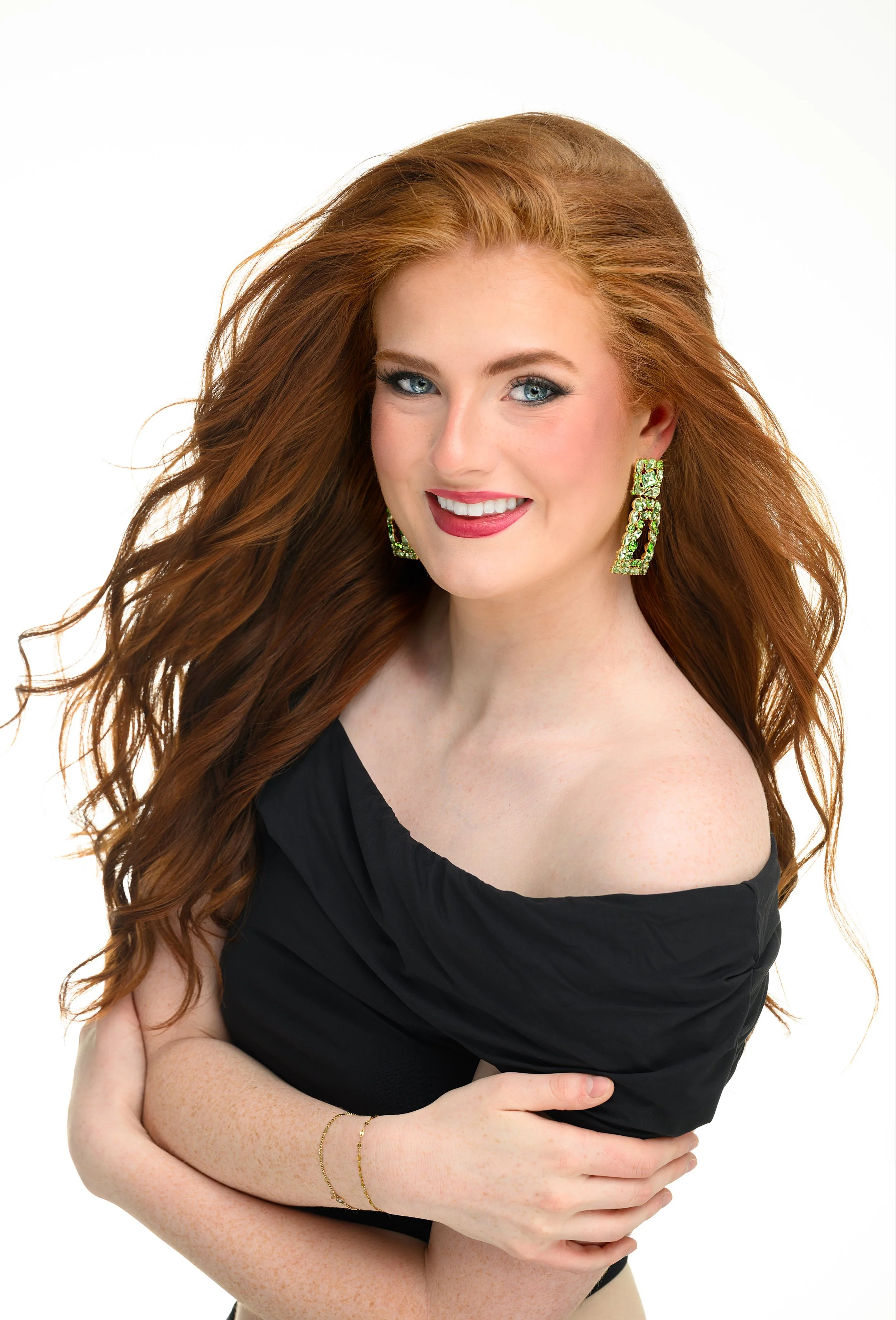 A woman with long, wavy red hair and blue eyes smiling at the camera. She is wearing a black top with a gold ring detail.