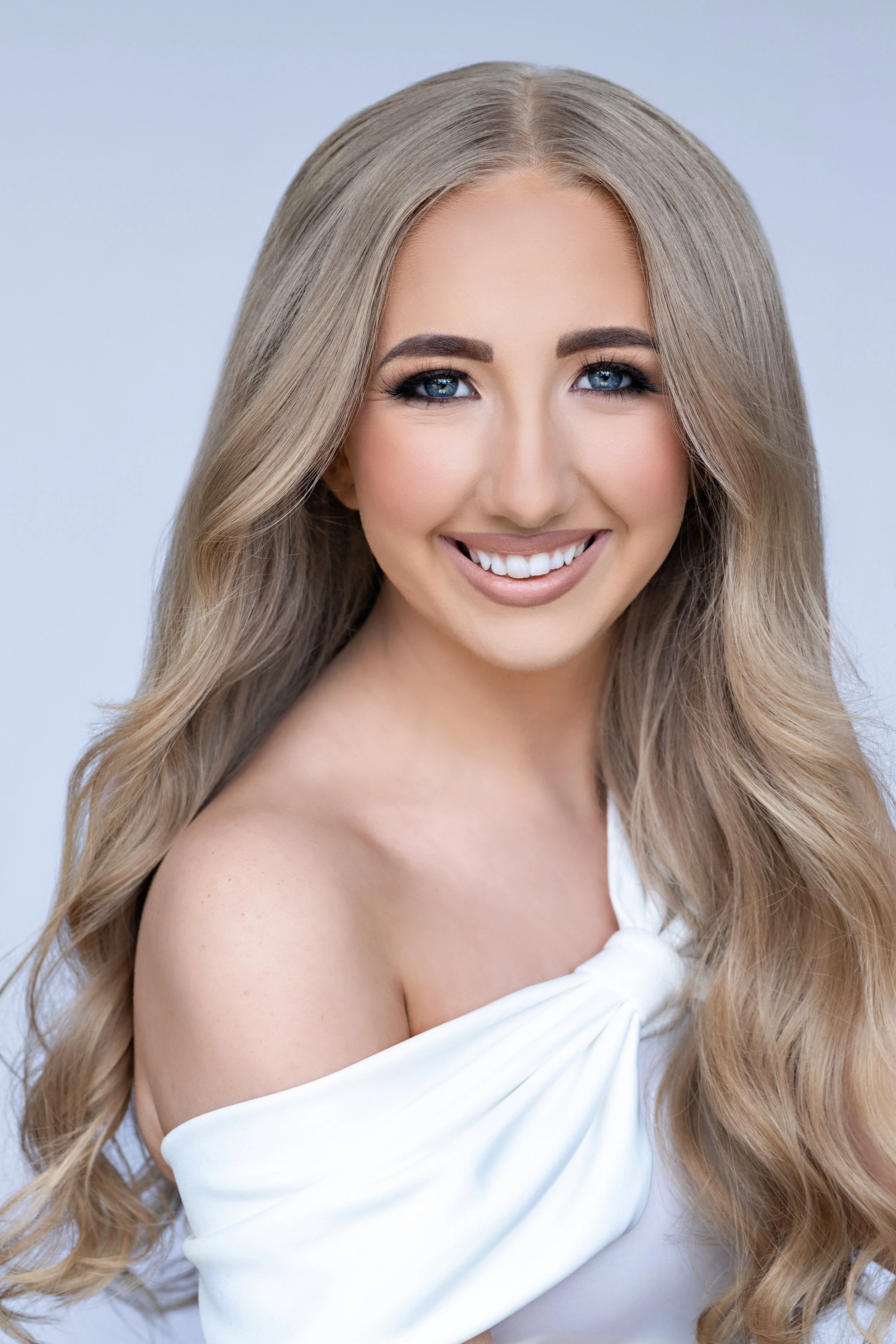 Smiling woman in a white top with lace trim, wearing gold earrings, with long wavy hair.