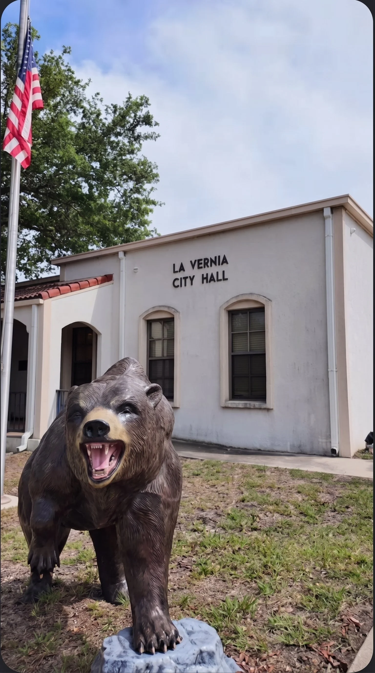 A bear sculpture with an open mouth, set in front of the La Vernia City Hall building, with trees, American flag, and cloudy sky in the background.