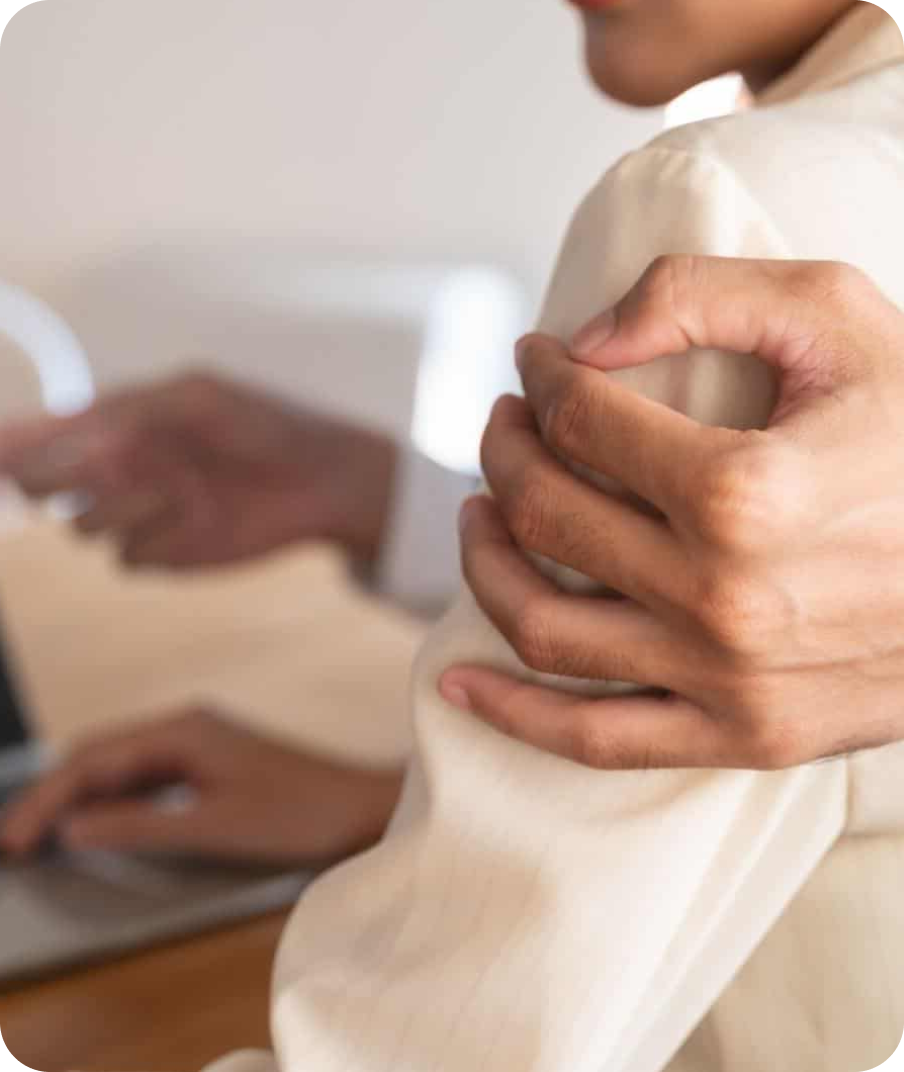 A person appears to be experiencing shoulder pain or discomfort, with another person in the background working on a laptop.