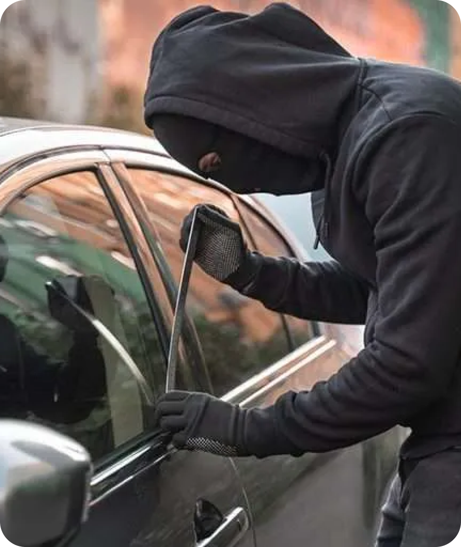 A person in black clothing and a ski mask attempting to break into a car using a metal tool.