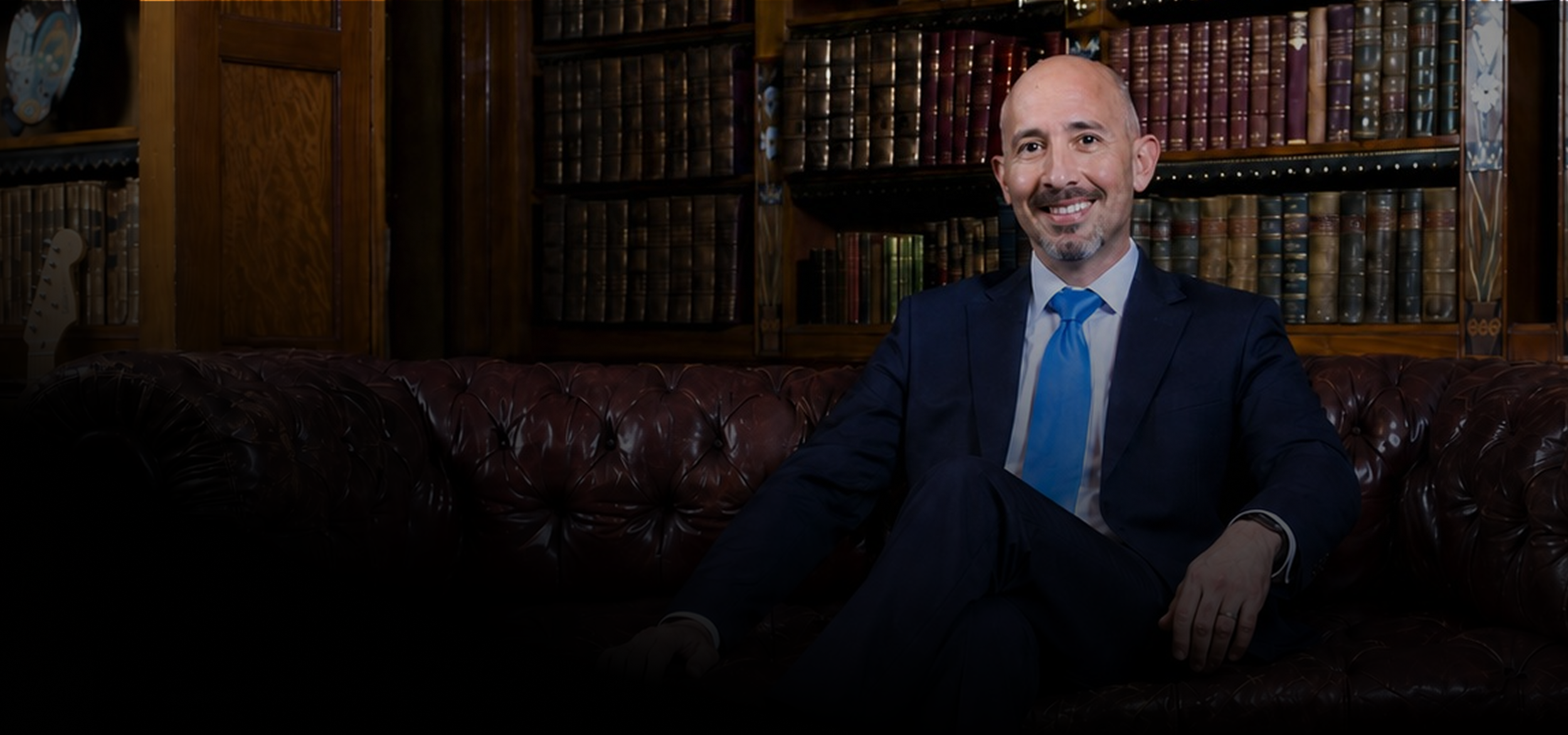 A man in a navy suit and blue tie sitting on a brown leather Chesterfield sofa in a wood-paneled library with bookshelves filled with leather-bound books in the background.