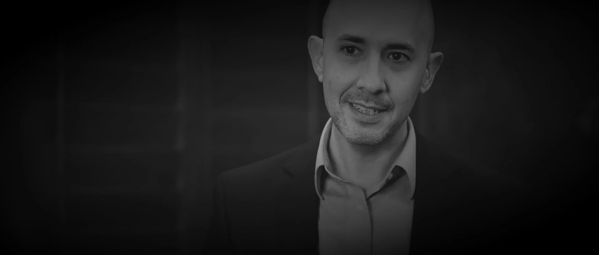 Black and white image of a man with short hair, wearing a suit and an open-collared shirt, smiling slightly while looking to the side.