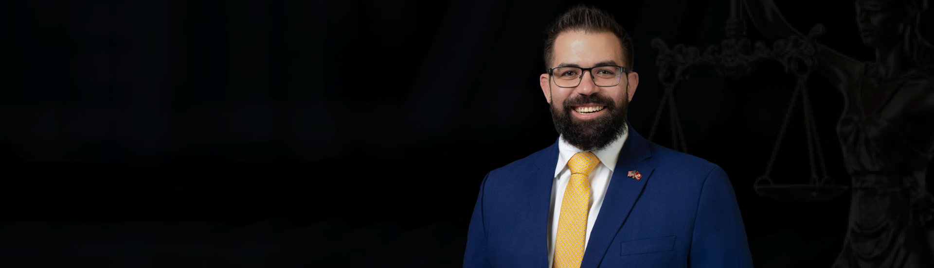 A man with glasses, a beard, and a big smile, wearing a blue suit, white shirt, yellow tie, and a small American flag pin, standing in front of a dark background with a faint image of Lady Justice holding scales.