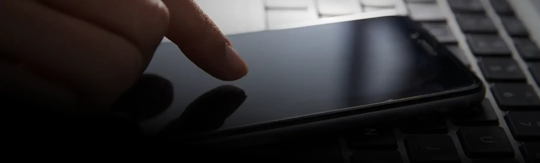 A person's finger touching the screen of a mobile phone placed on a laptop keyboard.