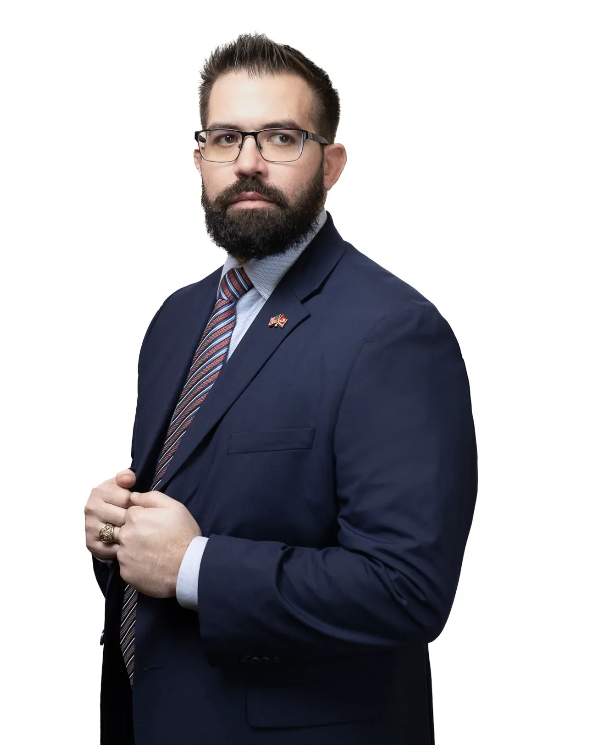 A man with a beard and glasses wearing a navy suit, light blue shirt, striped tie, and small American flag pin on lapel, posing confidently against a white background.