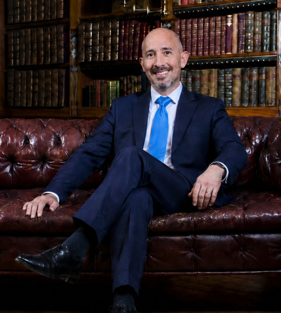 A man in a navy suit, light blue tie, and white shirt sitting on a brown leather sofa in a room filled with wooden bookshelves containing old, colorful books. He is smiling, with one leg crossed over the other.