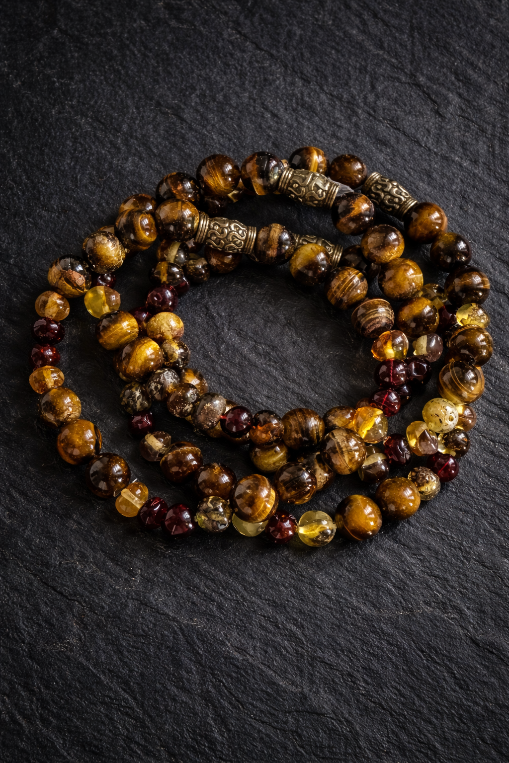 A beaded necklace made of round, polished stones in brown, amber, yellow, and red tones with ornate metallic spacers, resting on a black textured surface.