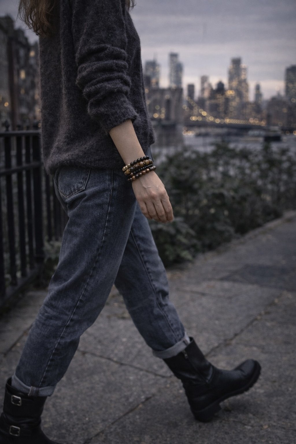 A person walking outdoors at dusk in an urban setting, wearing a dark sweater, jeans, and black boots, with a city skyline and river in the background.