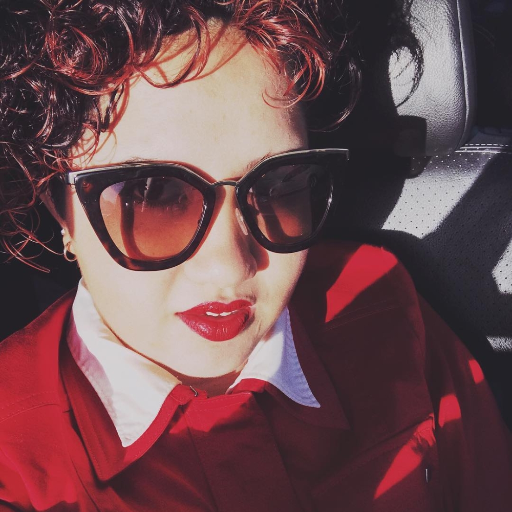 Selfie de Bella, assise sur un siège de voiture, cadrée en plan poitrine. Elle a les cheveux roux, du rouge à lèvres, et porte une chemise rouge. Elle a des lunettes de soleil dans lesquelles on voit qu’elle fait un clin d’œil.
