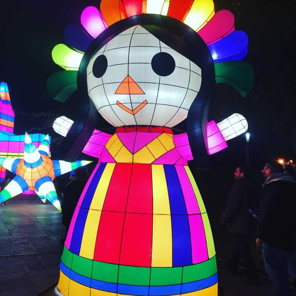 Photographie prise de nuit dans une rue, montrant des installations d’art lumineuses et colorées de quelques mètres de hauteur qui représentent des symboles culturels du Mexique, notamment au premier plan une poupée de chiffon « Maria ».
