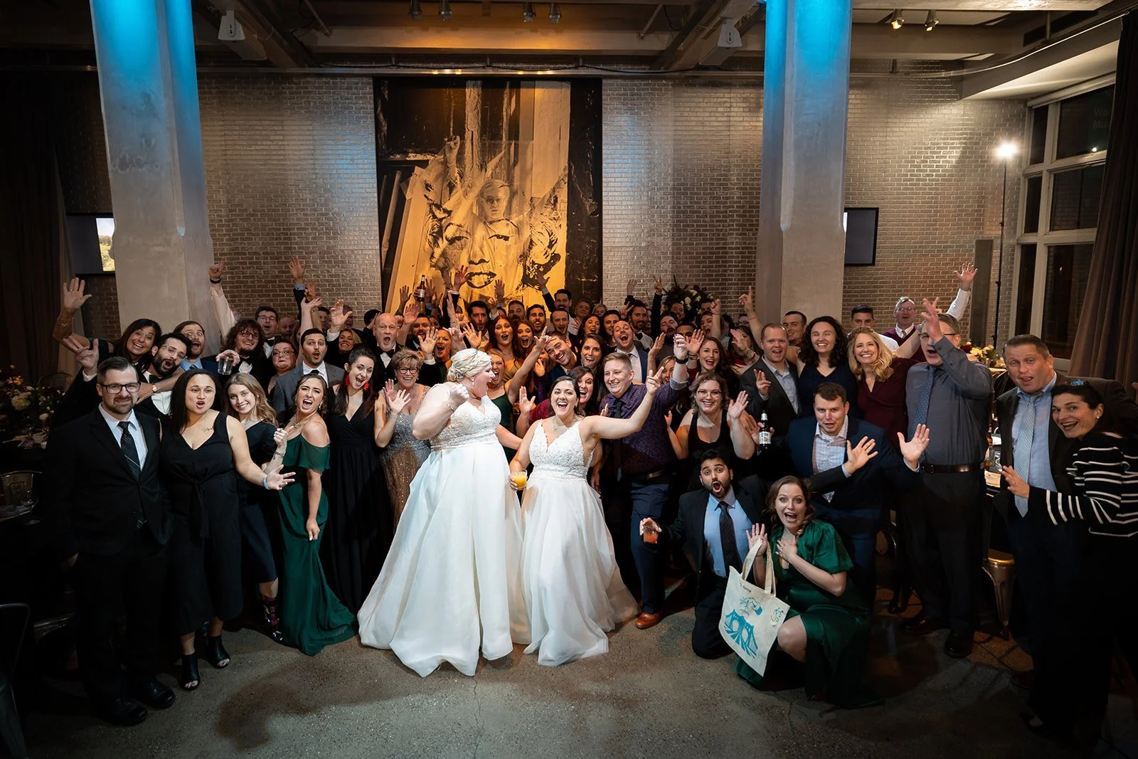 A large group of people celebrating at a wedding reception, including two women in white wedding dresses at the center and many others in formal attire, smiling and raising their hands.
