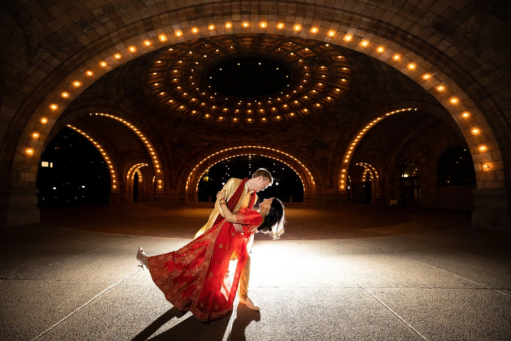 A couple dressed in traditional Indian attire sharing a dance under a large, ornate, illuminated arches and domed ceiling at night.