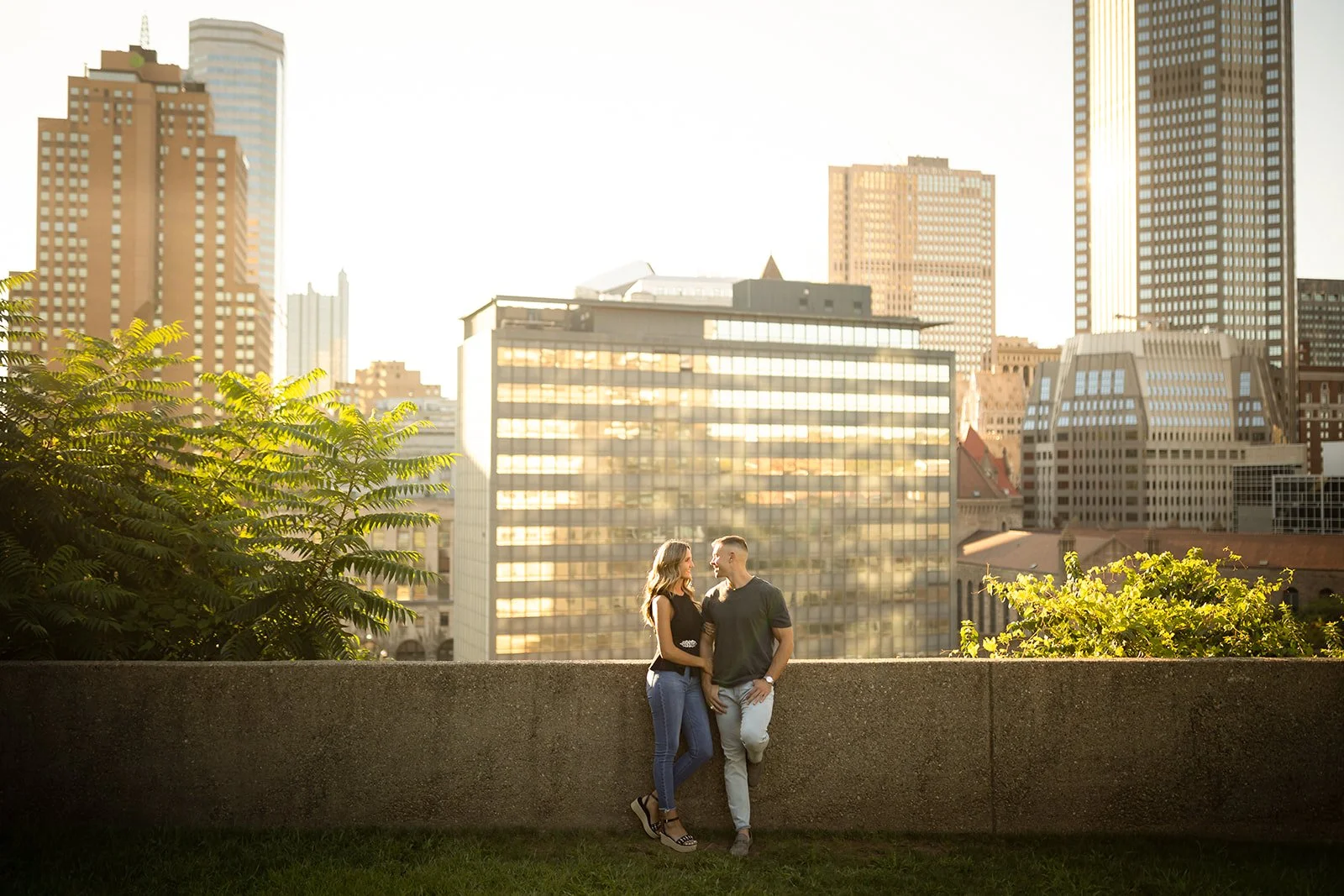 A couple standing close together on a rooftop ledge in front of a city skyline at sunset, with tall buildings and greenery around.