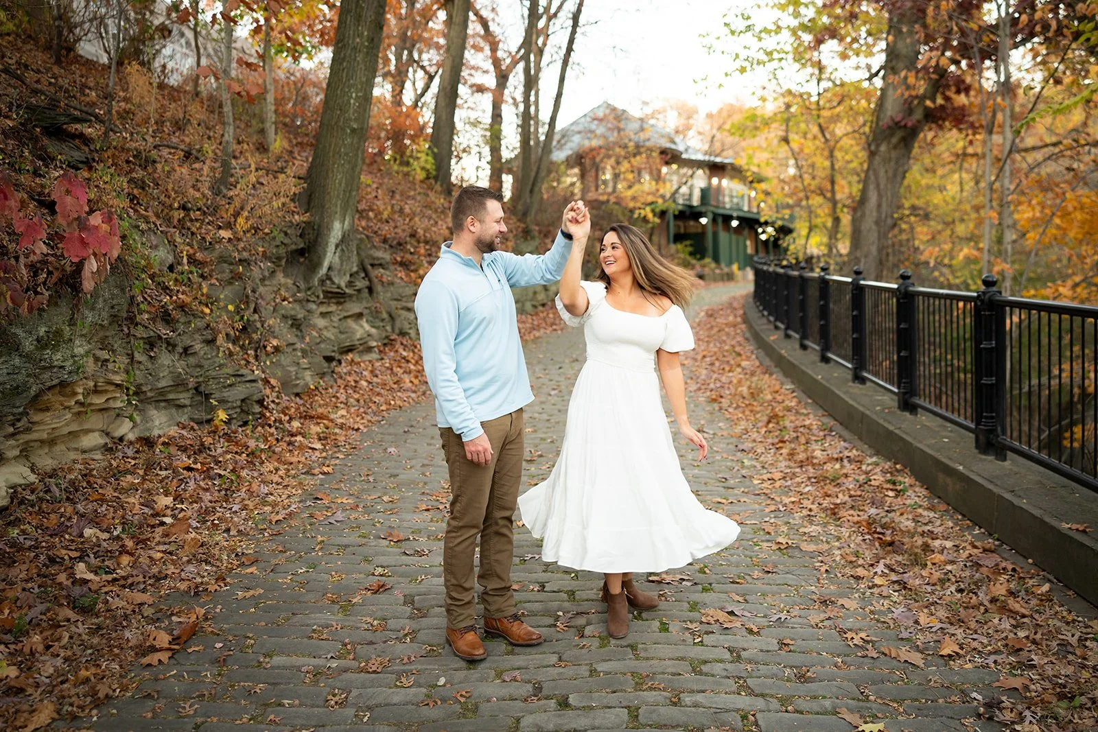 A man and woman dancing together on a autumnal walkway with fallen leaves, trees with orange and yellow leaves, a black railing on the right, and a house in the background.