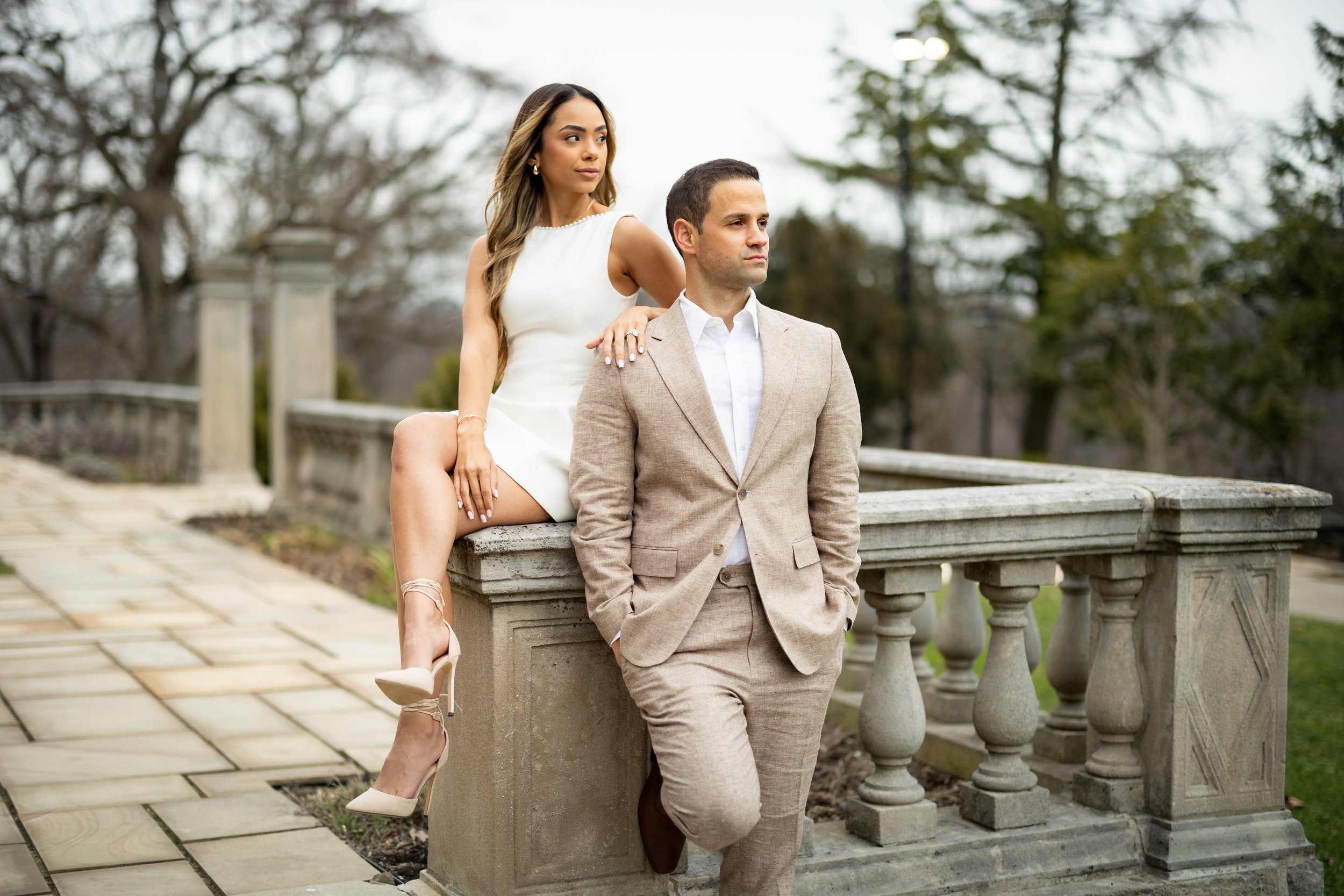 A man in a beige suit with his hands in his pockets and a woman in a white dress and heels sitting on a stone railing outdoors. The woman is resting her hand on the man's shoulder and looking to the side. The background shows trees and a cloudy sky.