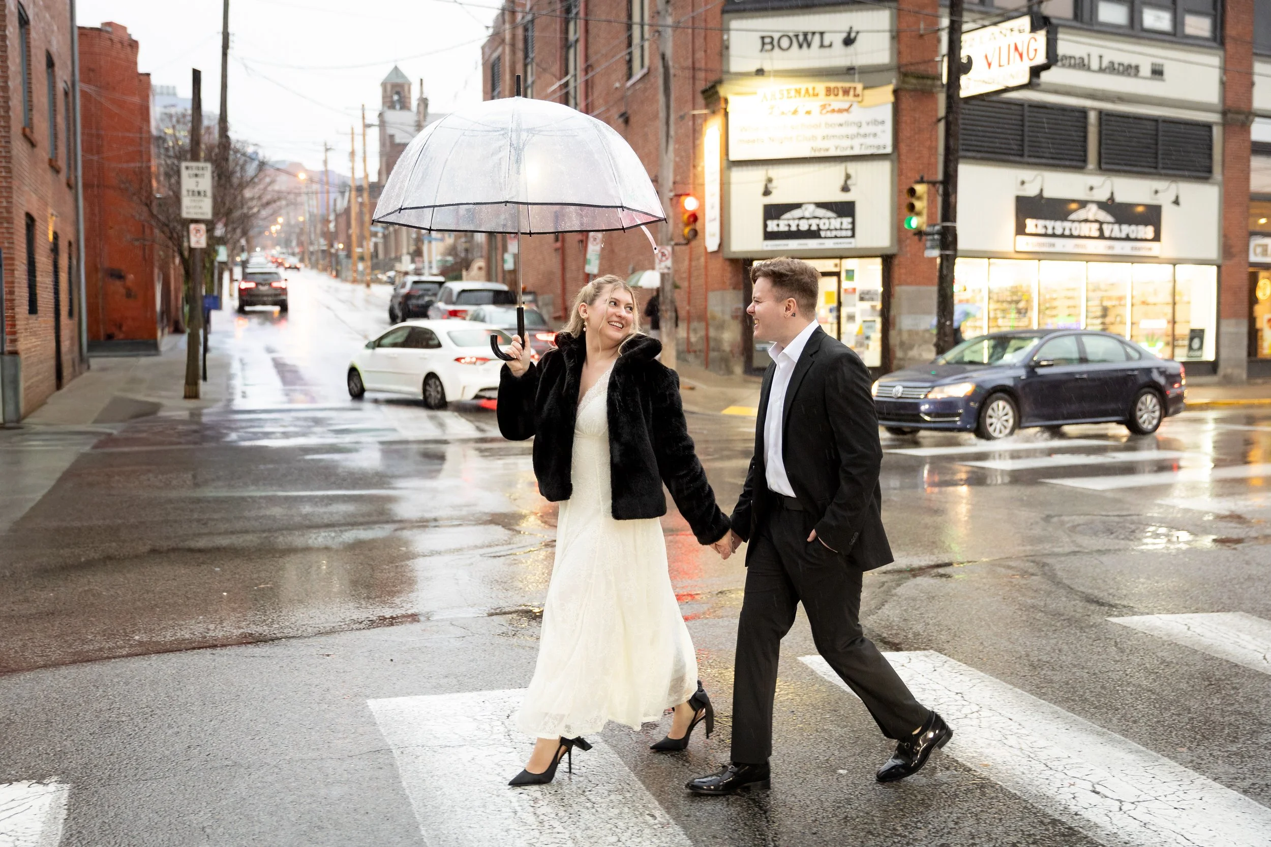 A couple dressed in formal attire walking hand-in-hand across a rainy city street while sharing an umbrella, with wet pavement, cars, and brick buildings in the background.