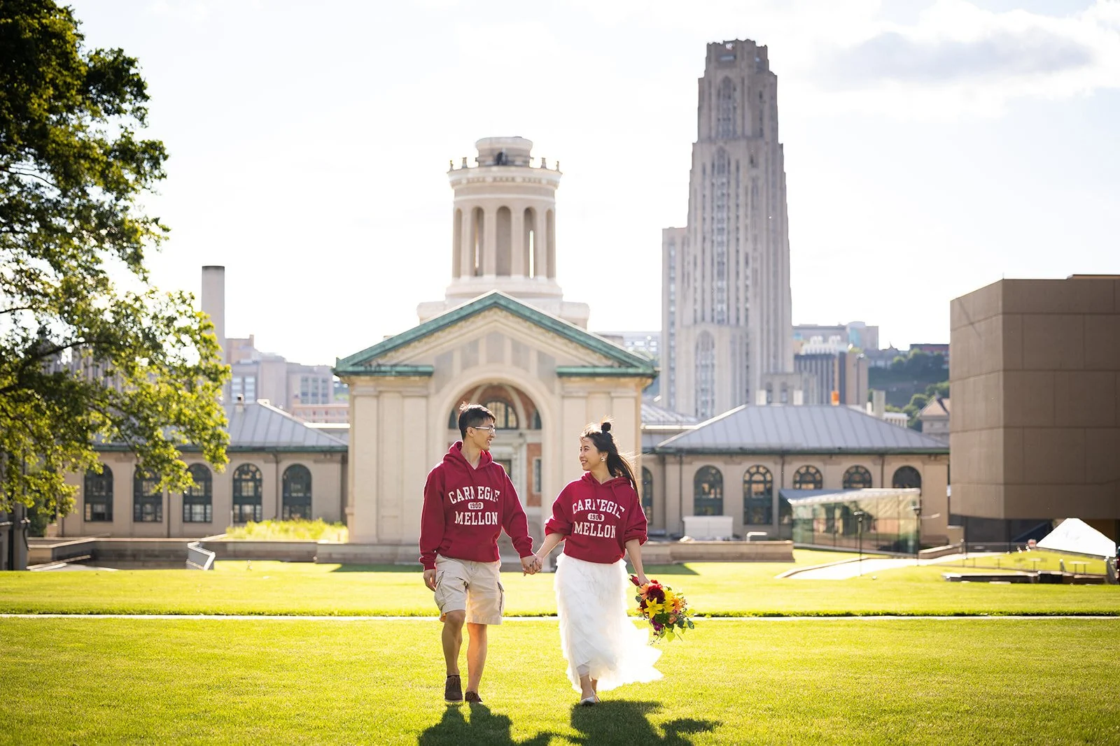 A young couple holding hands and walking on a grassy field in front of a historic building. The woman is holding a bouquet of colorful flowers, and both are wearing red sweatshirts with 'Carnegie Mellon' written on them. The background features a city skyline with tall buildings and a bright, sunny sky.