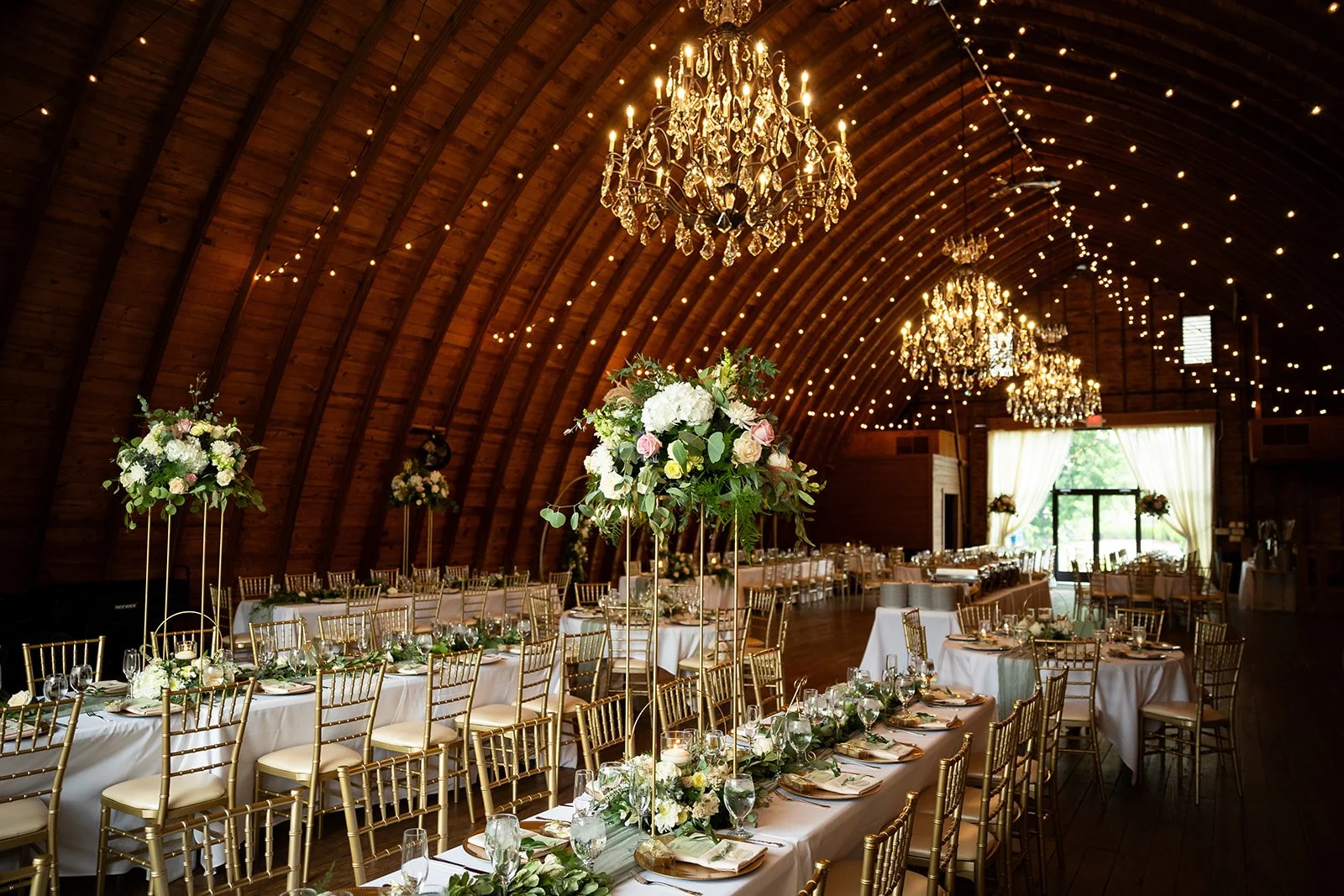 Elegant indoor wedding reception with long rectangular tables covered in white tablecloths, decorated with floral centerpieces featuring white and pink flowers and greenery, surrounded by gold chairs, under a wooden arched ceiling with chandeliers and string lights, with large windows and white curtains at the far end.