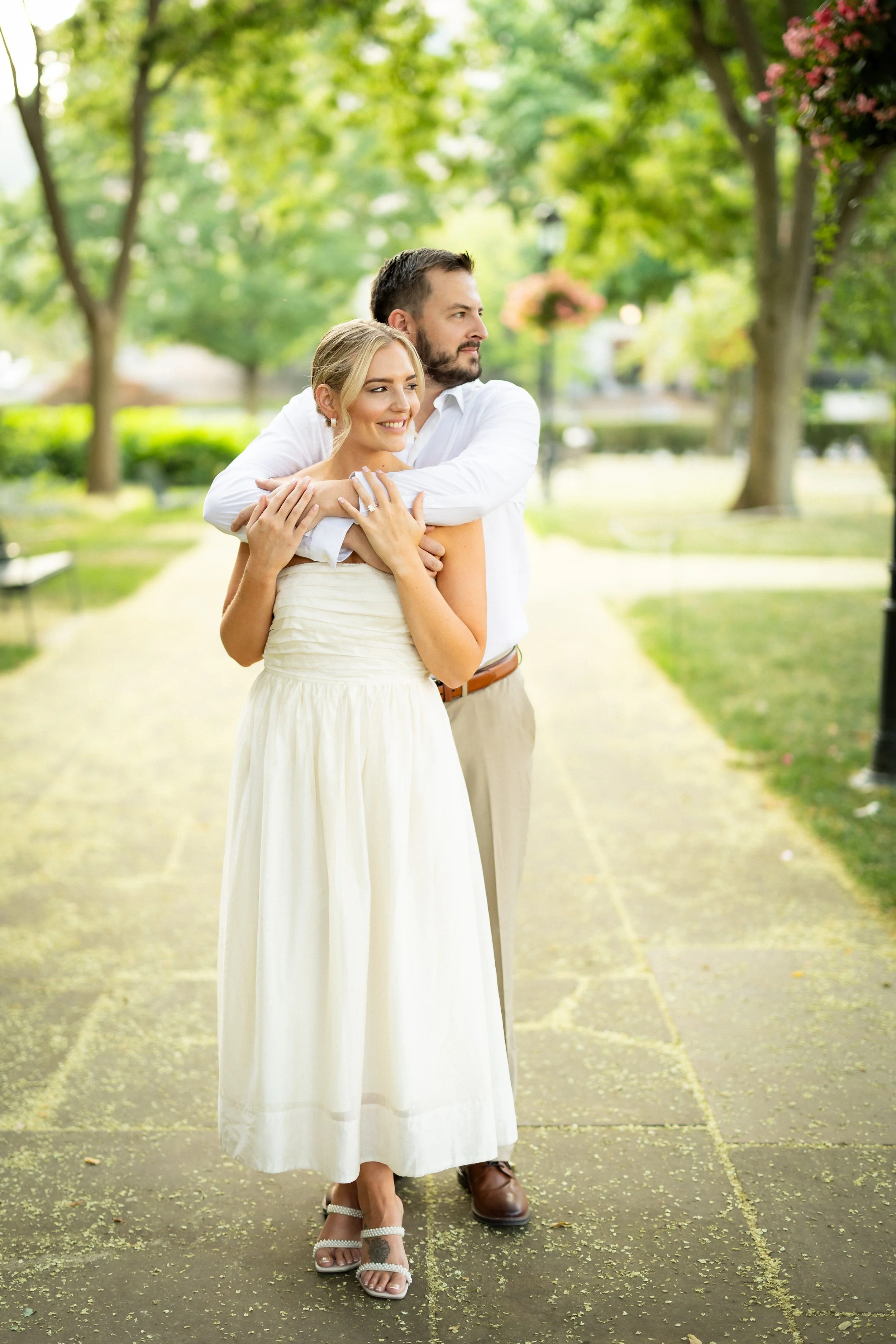 A smiling woman in a white dress and a man in light-colored pants and a white shirt embracing outdoors on a sunny day with green trees and a pathway in the background.