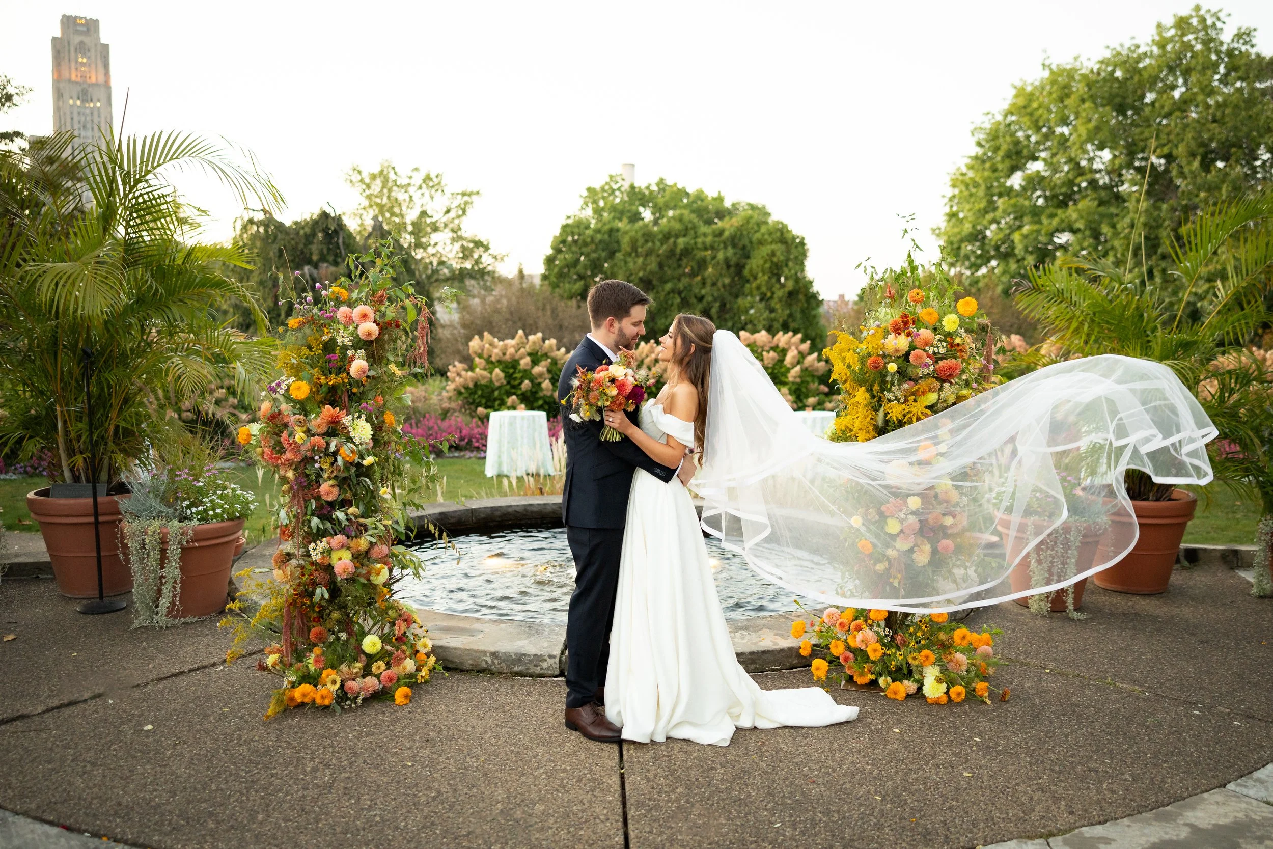 A bride and groom stand close together in a garden, surrounded by colorful floral arrangements, during their wedding ceremony. The bride is wearing a white wedding dress with an off-shoulder design and a flowing veil, and she holds a bouquet of flowers. The groom is dressed in a dark suit. They are smiling at each other near a small fountain, with lush green trees and flowers in the background.