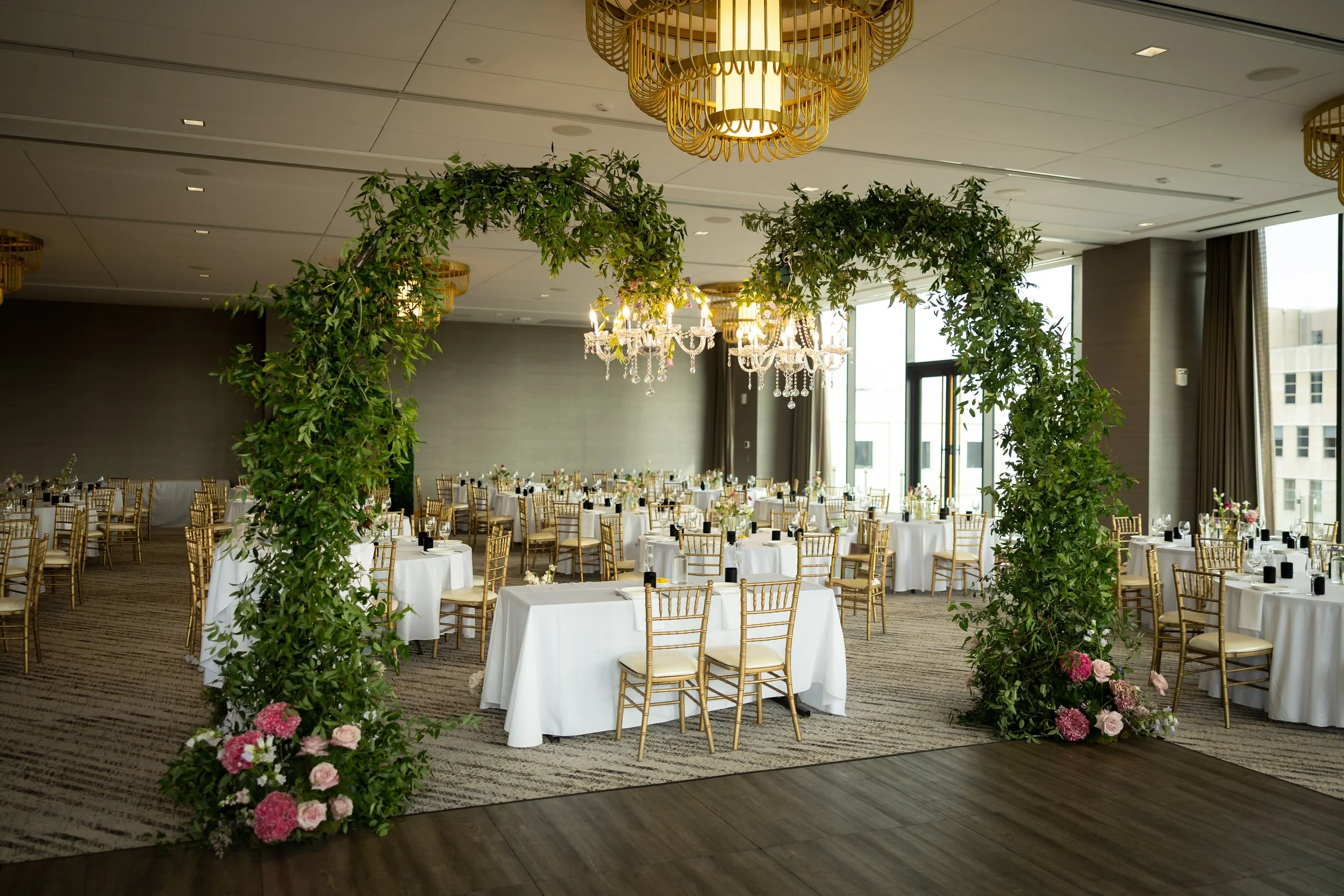 Elegant event space decorated with a floral arch and chandeliers, featuring round tables with white tablecloths and gold chairs, set up for a celebration.