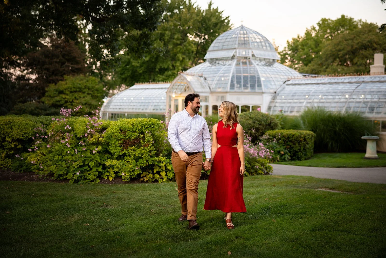 A couple walking hand in hand on grass in front of a greenhouse, with trees and pink flowers in the background.