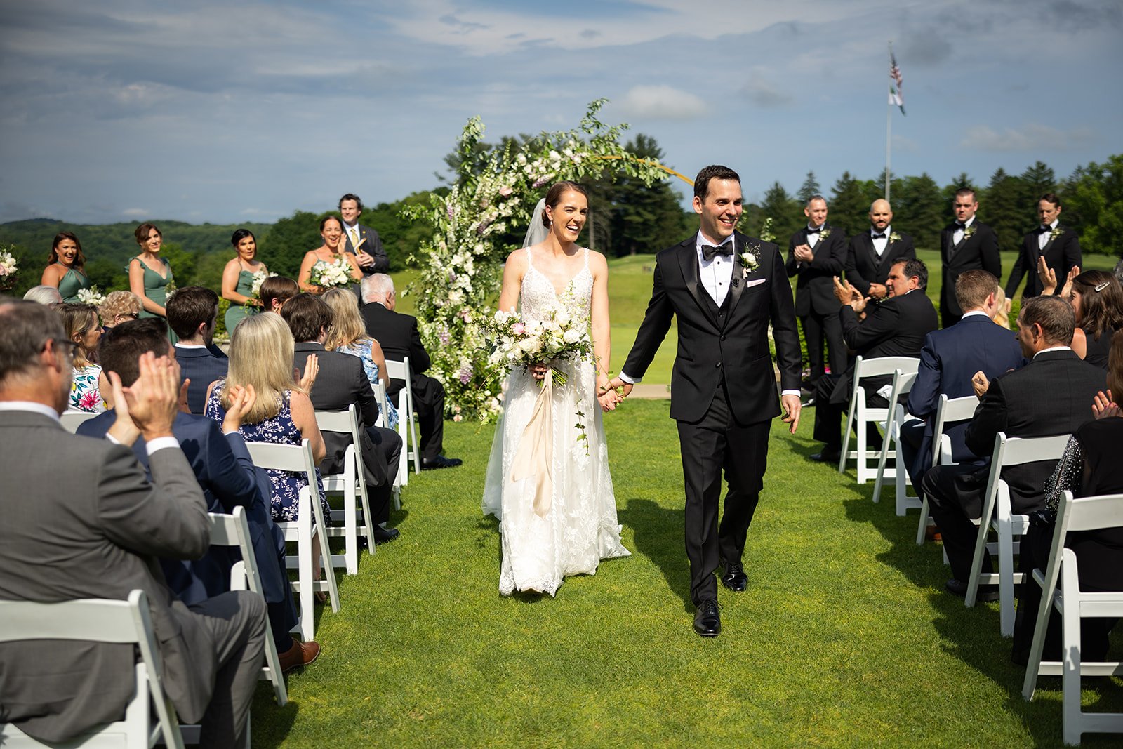 Bride and groom holding hands and walking down the aisle at an outdoor wedding ceremony, surrounded by seated guests and wedding party, with a floral arch and green landscape in the background.