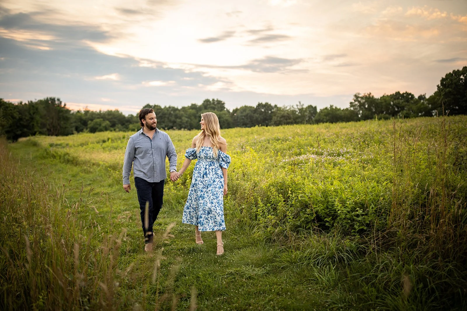 A man and woman holding hands and walking on a grassy path through a green field at sunset, smiling at each other.