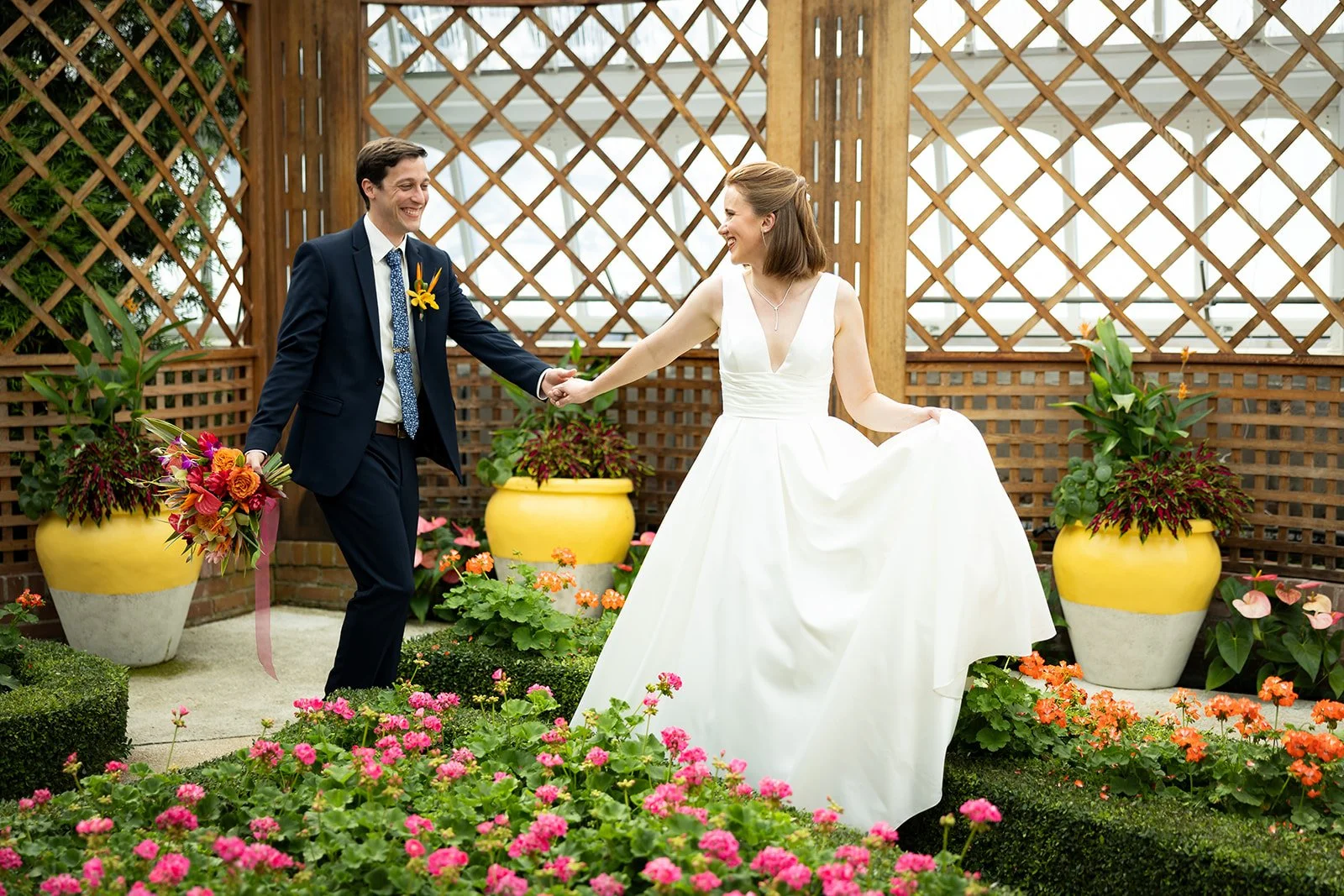 A bride and groom holding hands and smiling in a garden with colorful flowers and large yellow pots.
