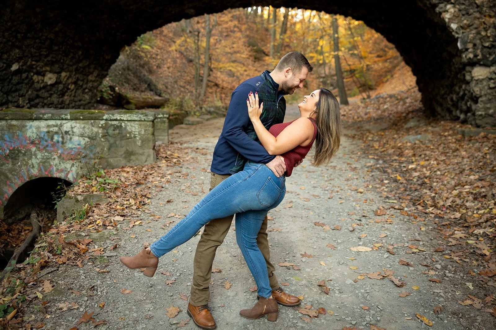 A couple dancing and smiling under a stone bridge in an autumn wooded area.
