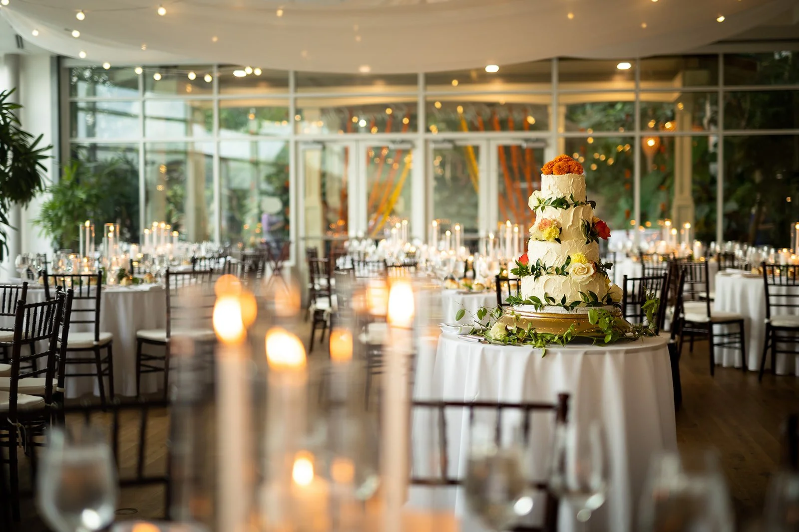 Elegant wedding reception hall with a large multi-tiered wedding cake decorated with flowers and greenery, surrounded by round tables with white tablecloths and chairs, illuminated by candles and string lights, with large glass windows in the background.