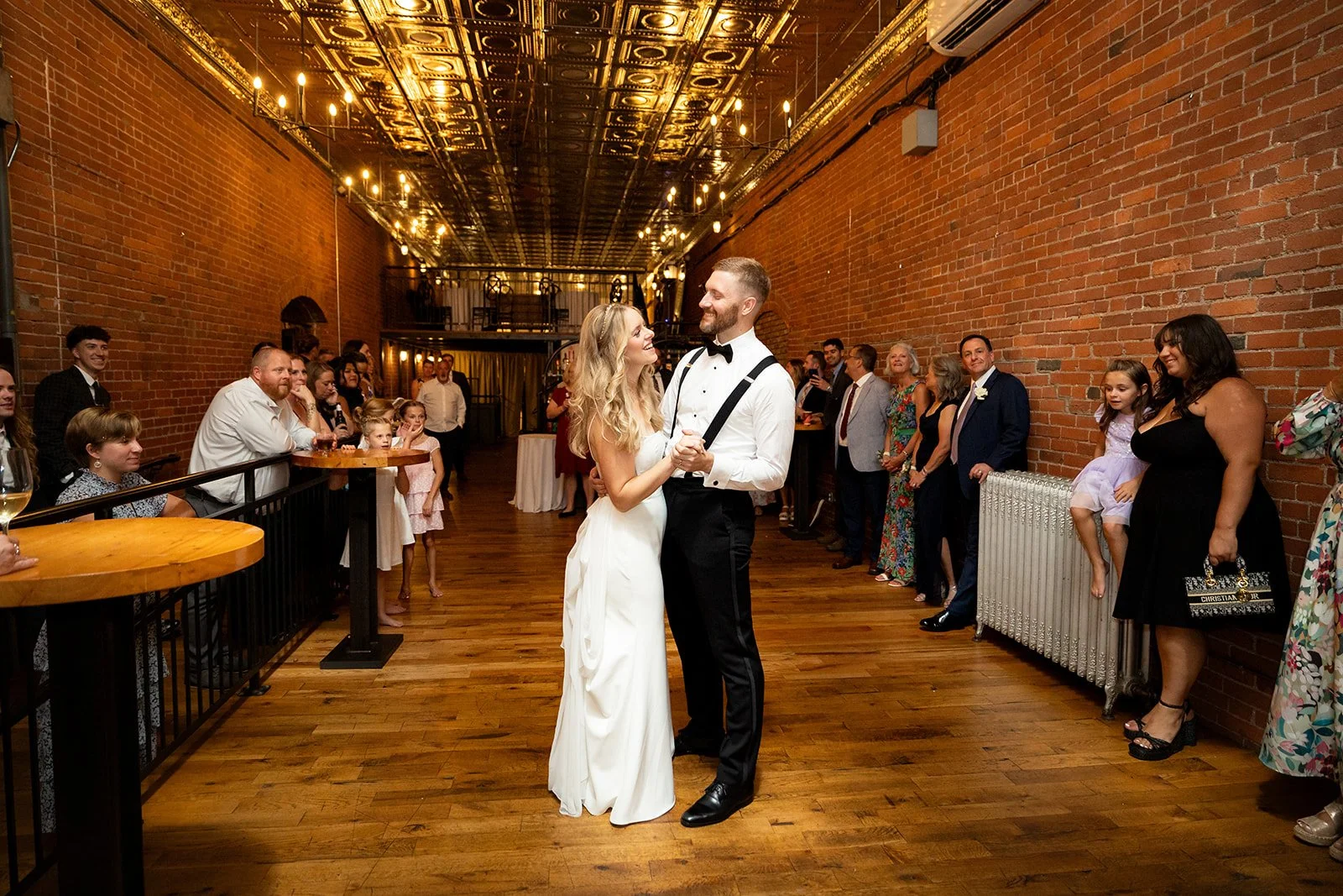 A bride and groom dancing at their wedding reception in a rustic, brick-walled venue with a vintage ceiling and a live band setup in the background. Guests are watching and smiling.
